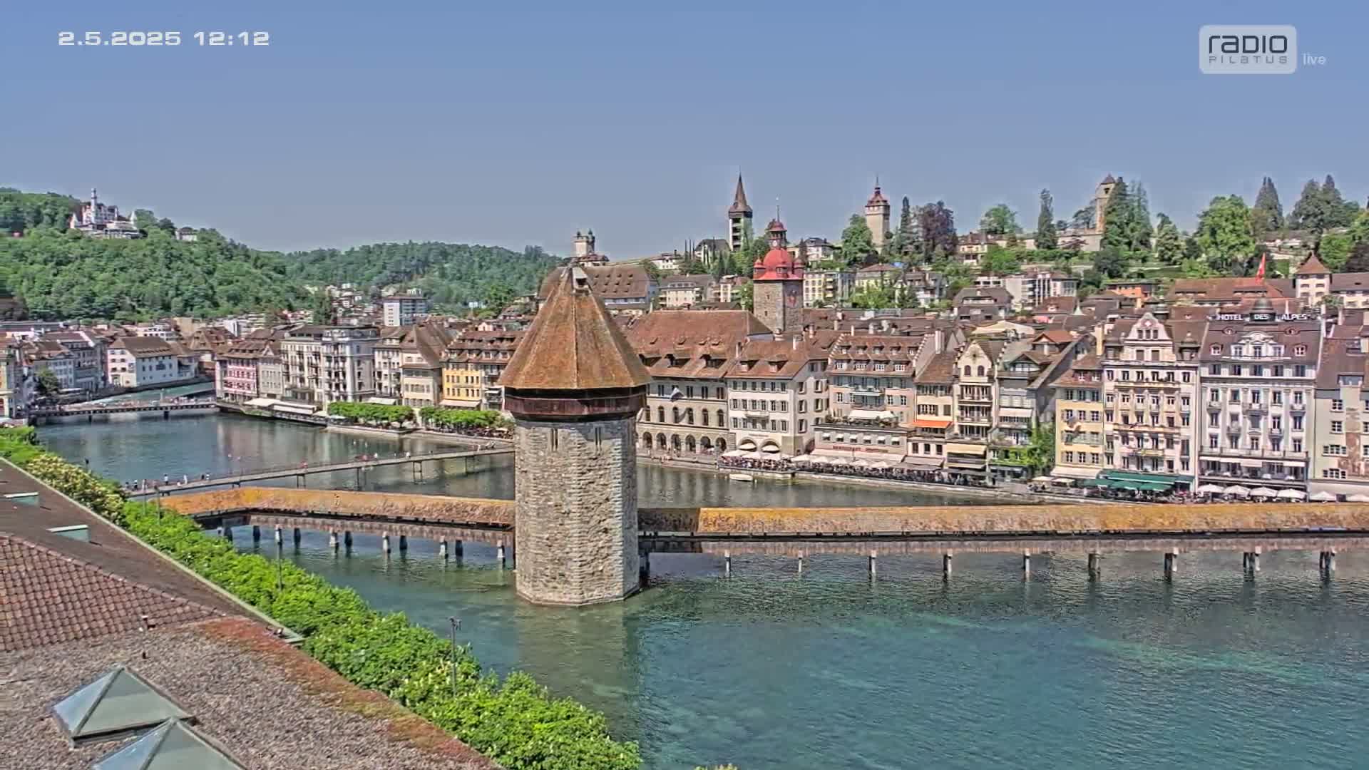 A stone tower stands in the middle of a covered wooden bridge spanning a clear body of water, with a town of colorful buildings and green hills in the background under a bright sunny sky.