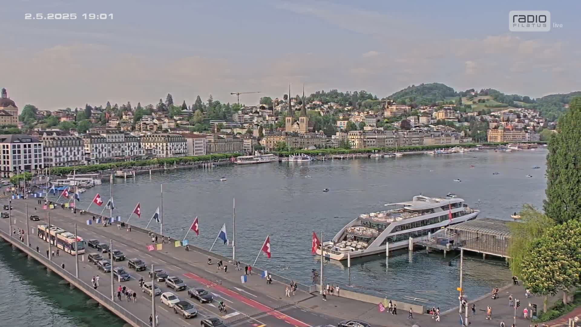 A waterfront city, under partly cloudy skies, is seen from above, showing a bridge with cars and pedestrians, boats on the water, and buildings along the shoreline.