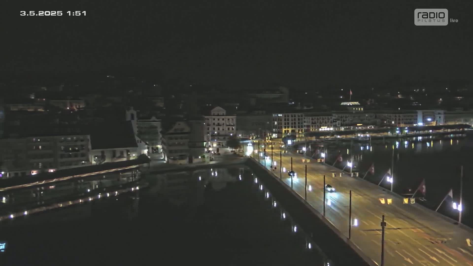 A nighttime aerial view shows a city's waterfront at night, with a bridge illuminated and a few vehicles traveling on it, reflecting in the calm water.
