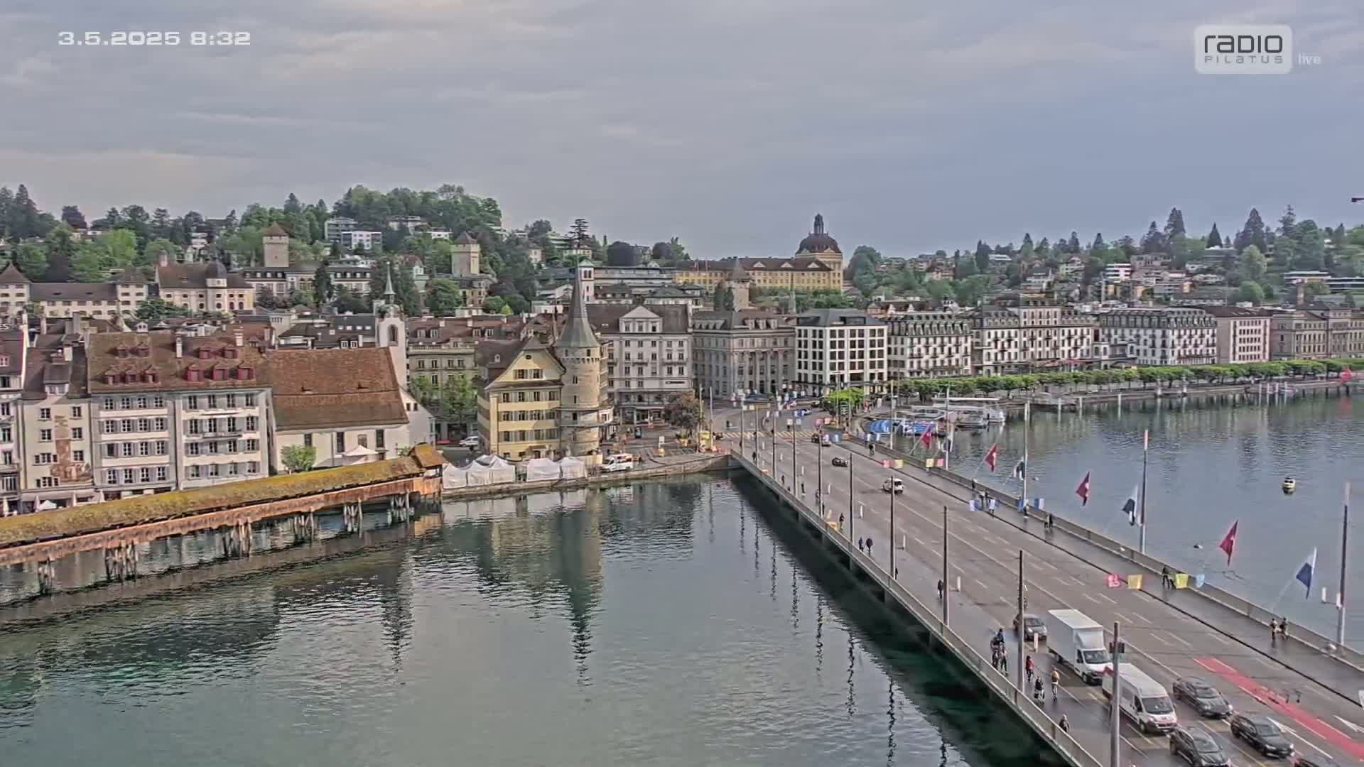A long bridge stretches across a calm body of water toward a European-style city under a cloudy sky.