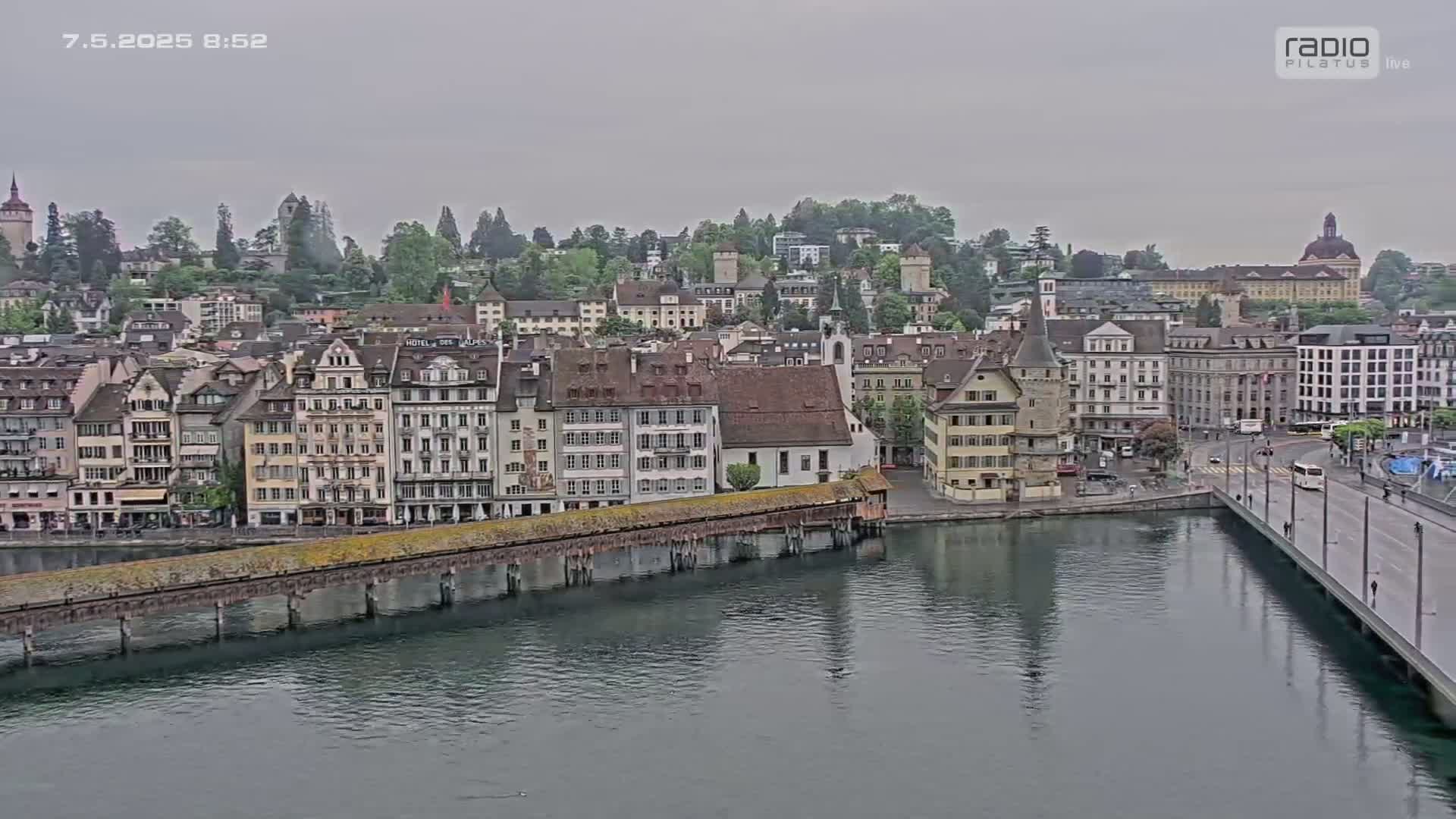 A covered bridge spans a calm river reflecting a cityscape of many buildings under an overcast sky.