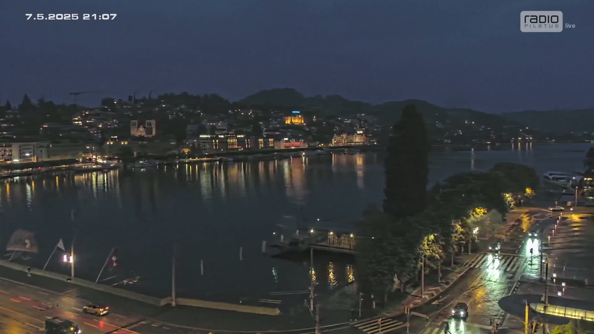A nighttime, rain-slicked city waterfront scene shows a calm body of water reflecting the lights of buildings on the opposite shore under a dark sky.