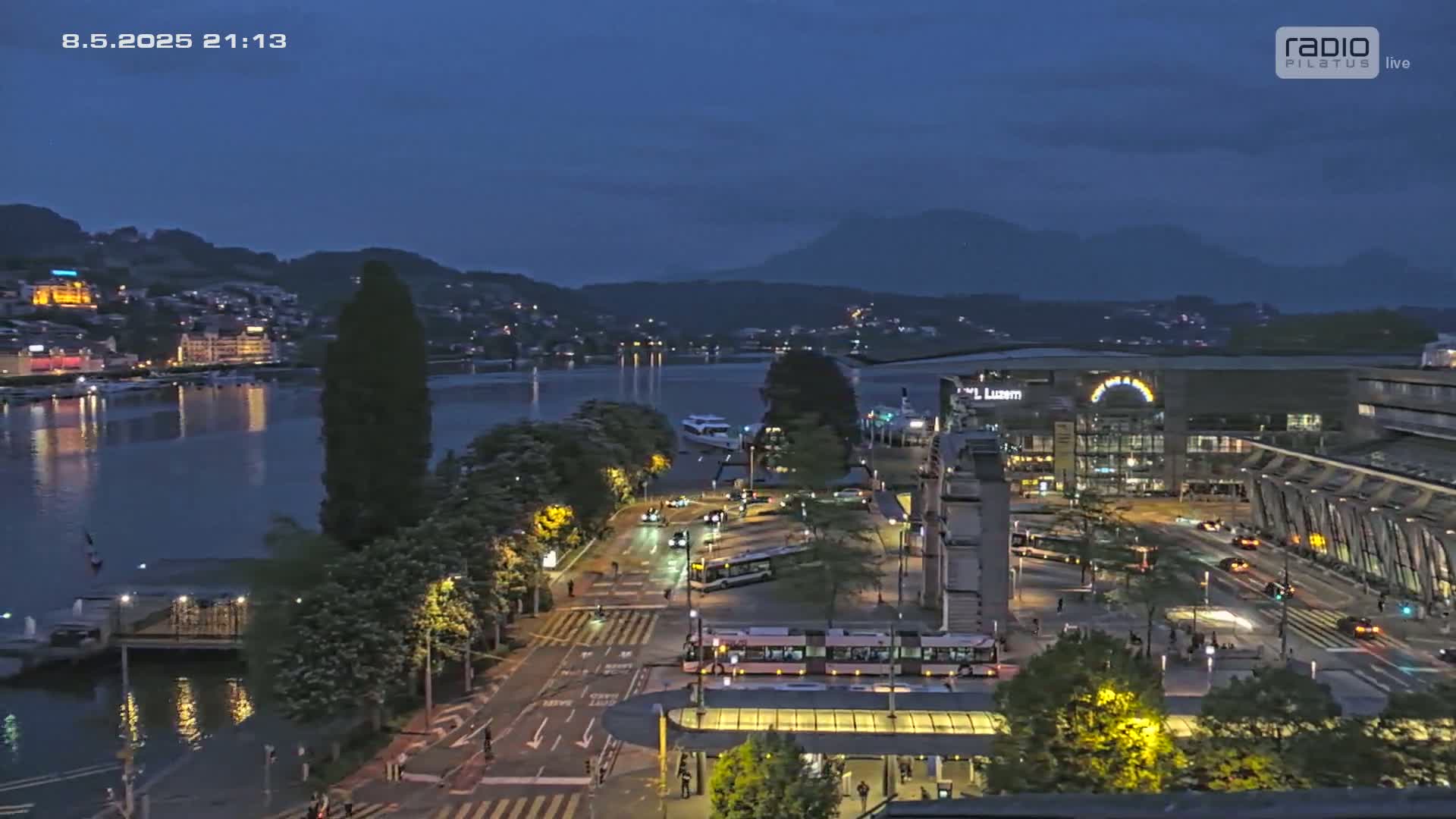 A nighttime view of a city waterfront with calm water reflecting lights, a street with trams, and buildings along a shoreline, under a dark blue twilight sky.