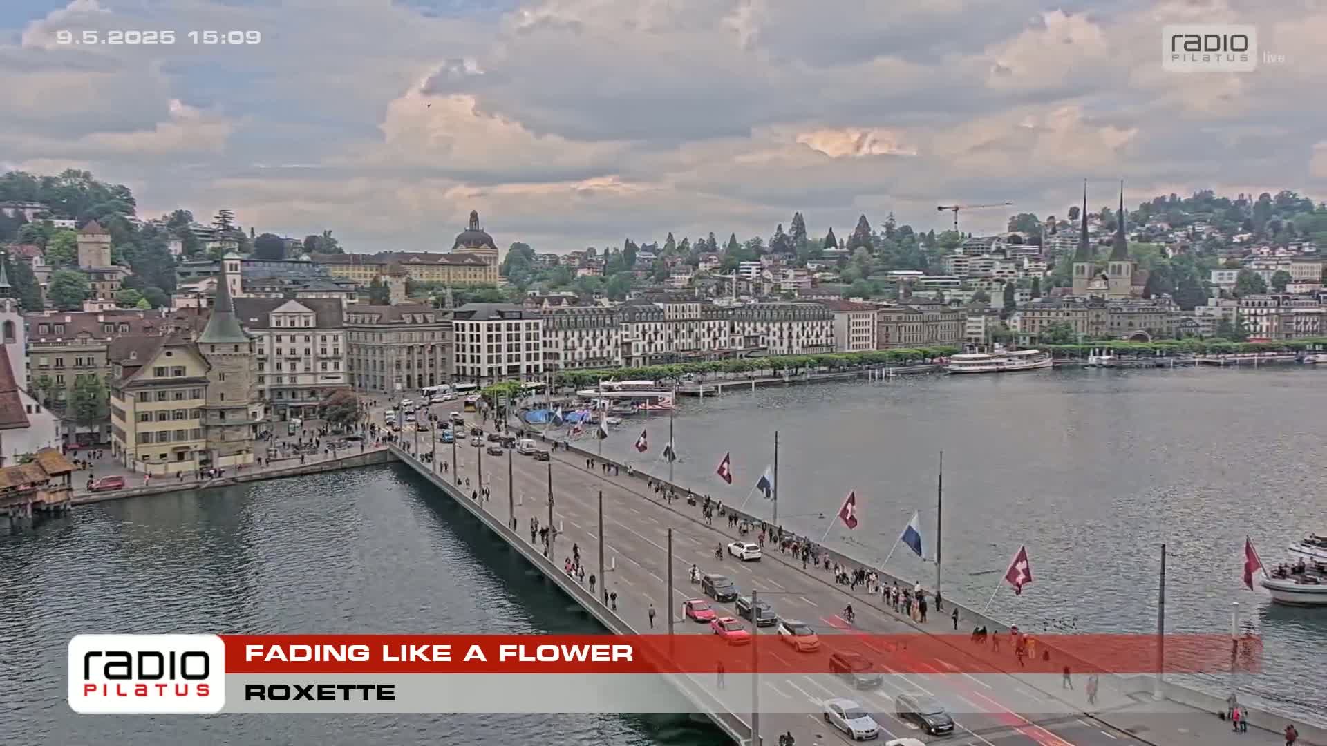 A partly cloudy day shows a city with many buildings along a body of water, a bridge with people walking and cars driving across it, and several Swiss flags along the bridge's sides.
