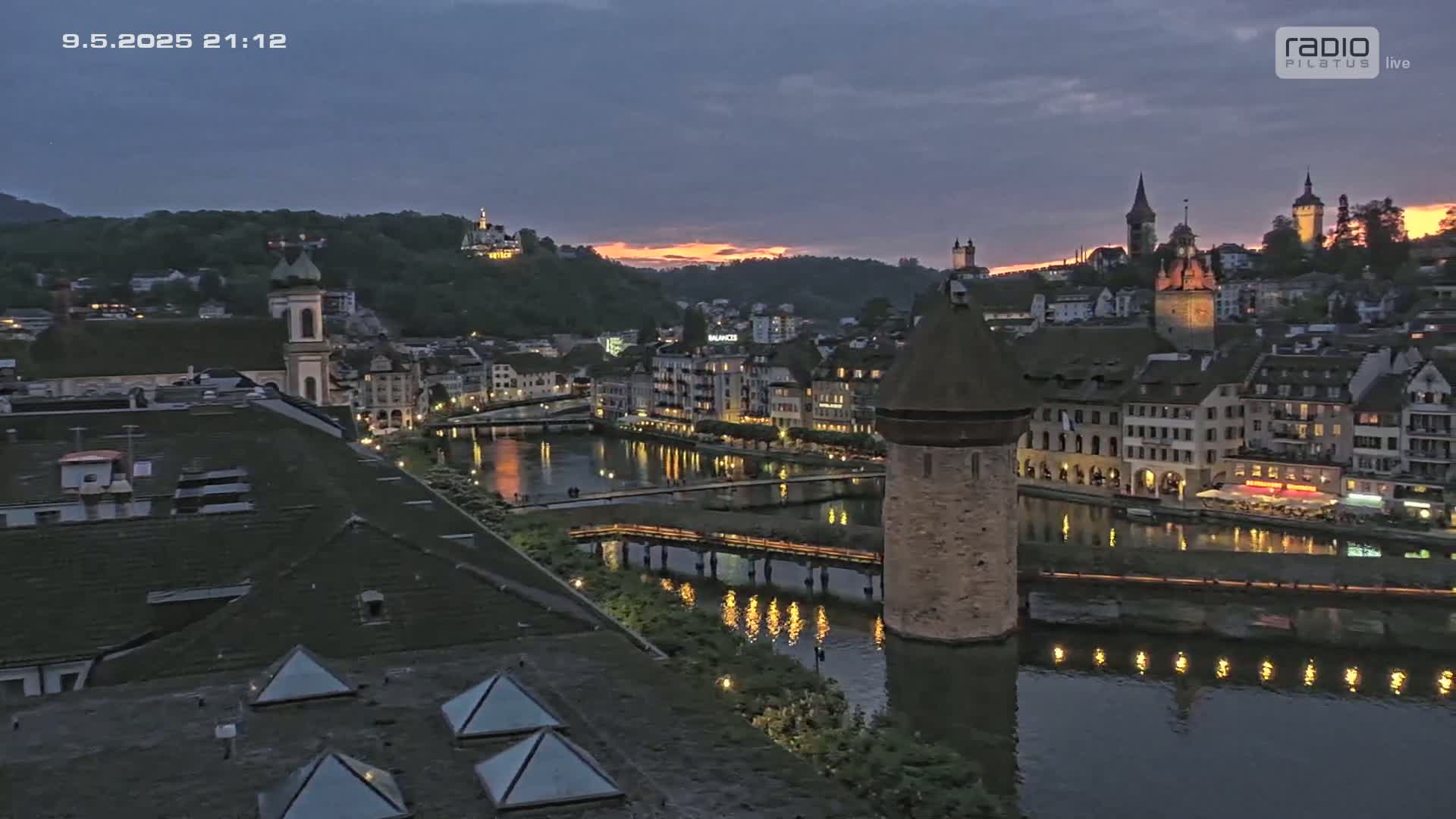 A twilight view of a European city with a river reflecting lights from buildings, a stone tower, and a bridge.