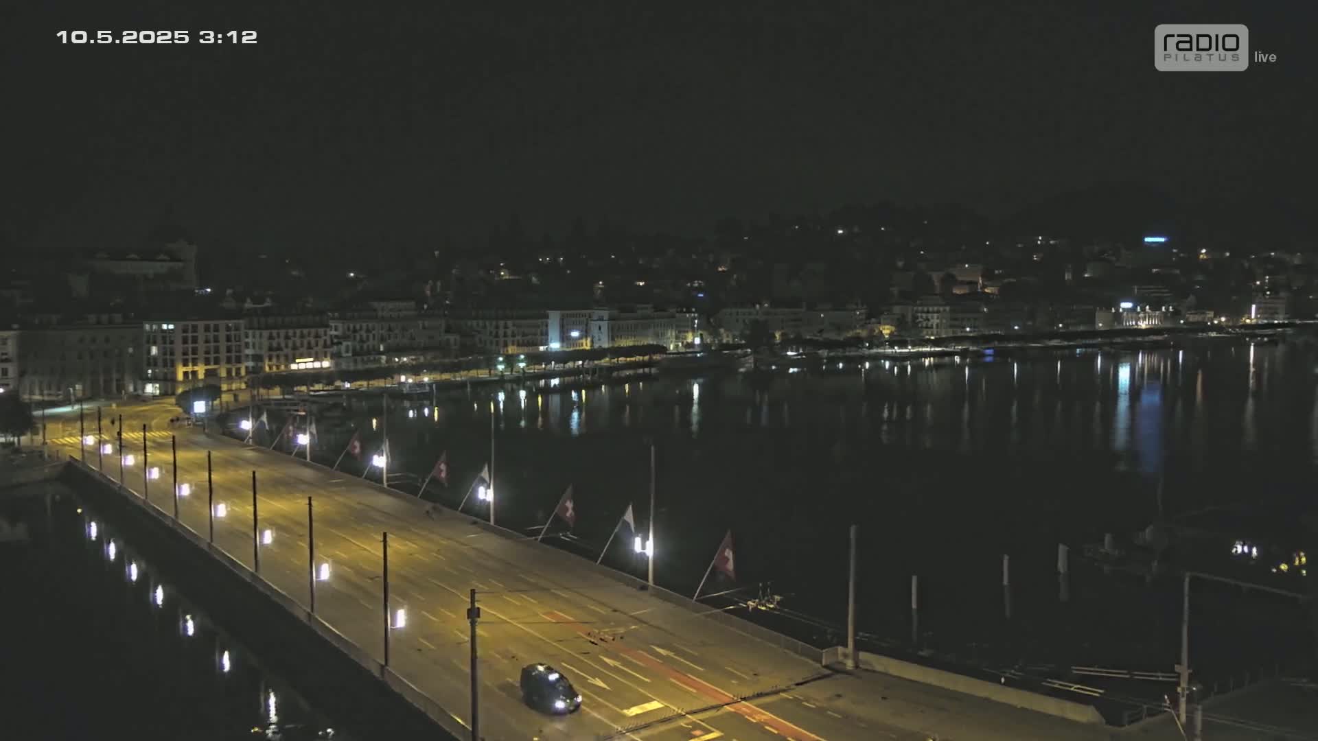 A nighttime, high-angle view shows a bridge illuminated with lights over dark water reflecting city lights, with a single vehicle driving across.