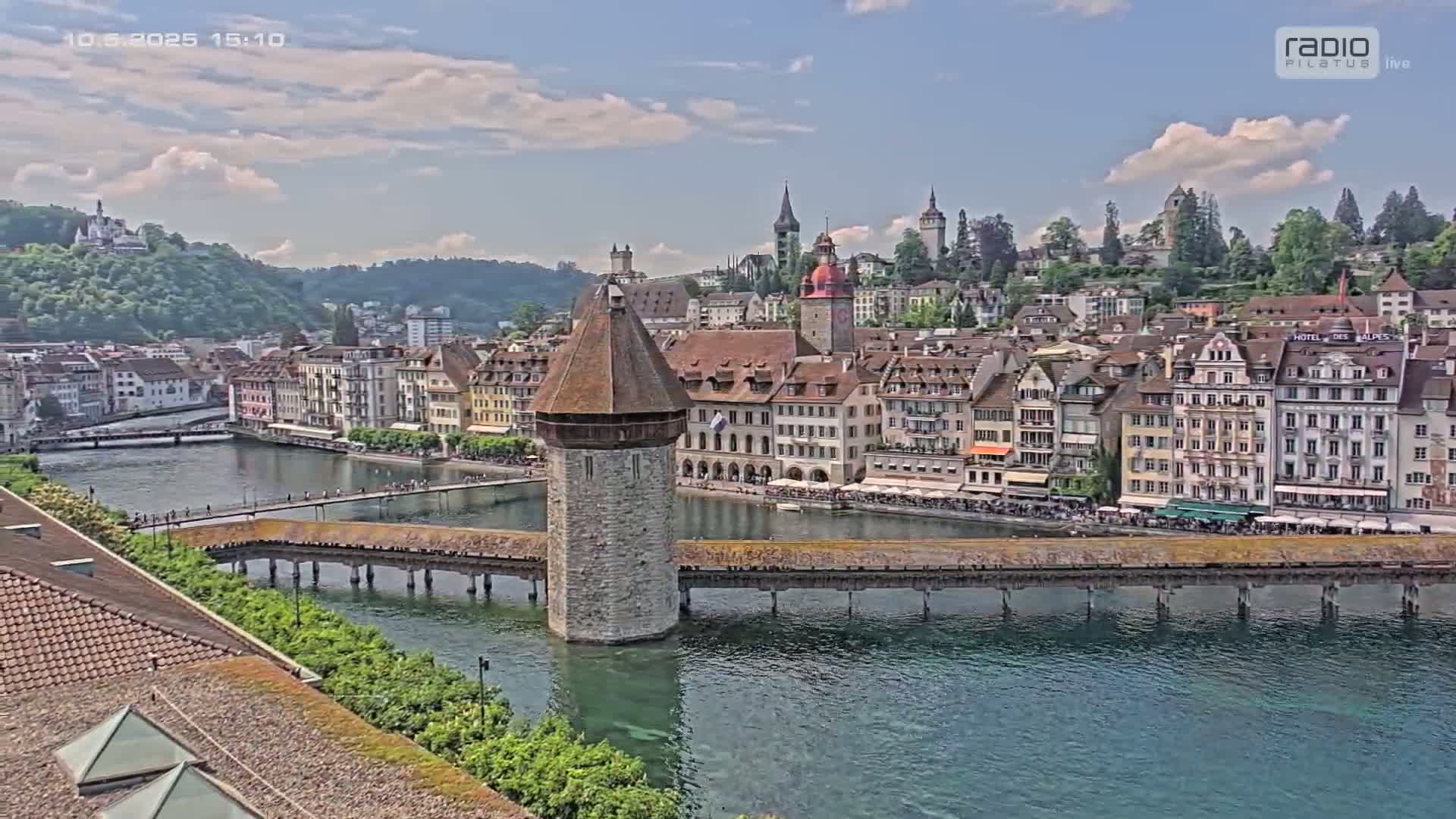 A stone tower stands in the middle of a wooden bridge spanning a river, with a charming town of colorful buildings lining the riverbanks under a partly cloudy sky.