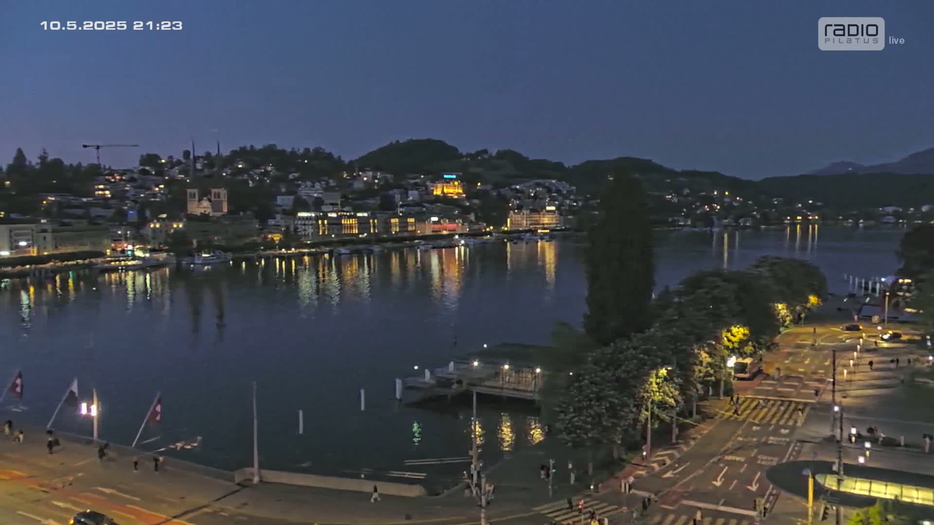 A nighttime view of a city on a lake, with buildings illuminated and a calm, dark body of water reflecting the lights.