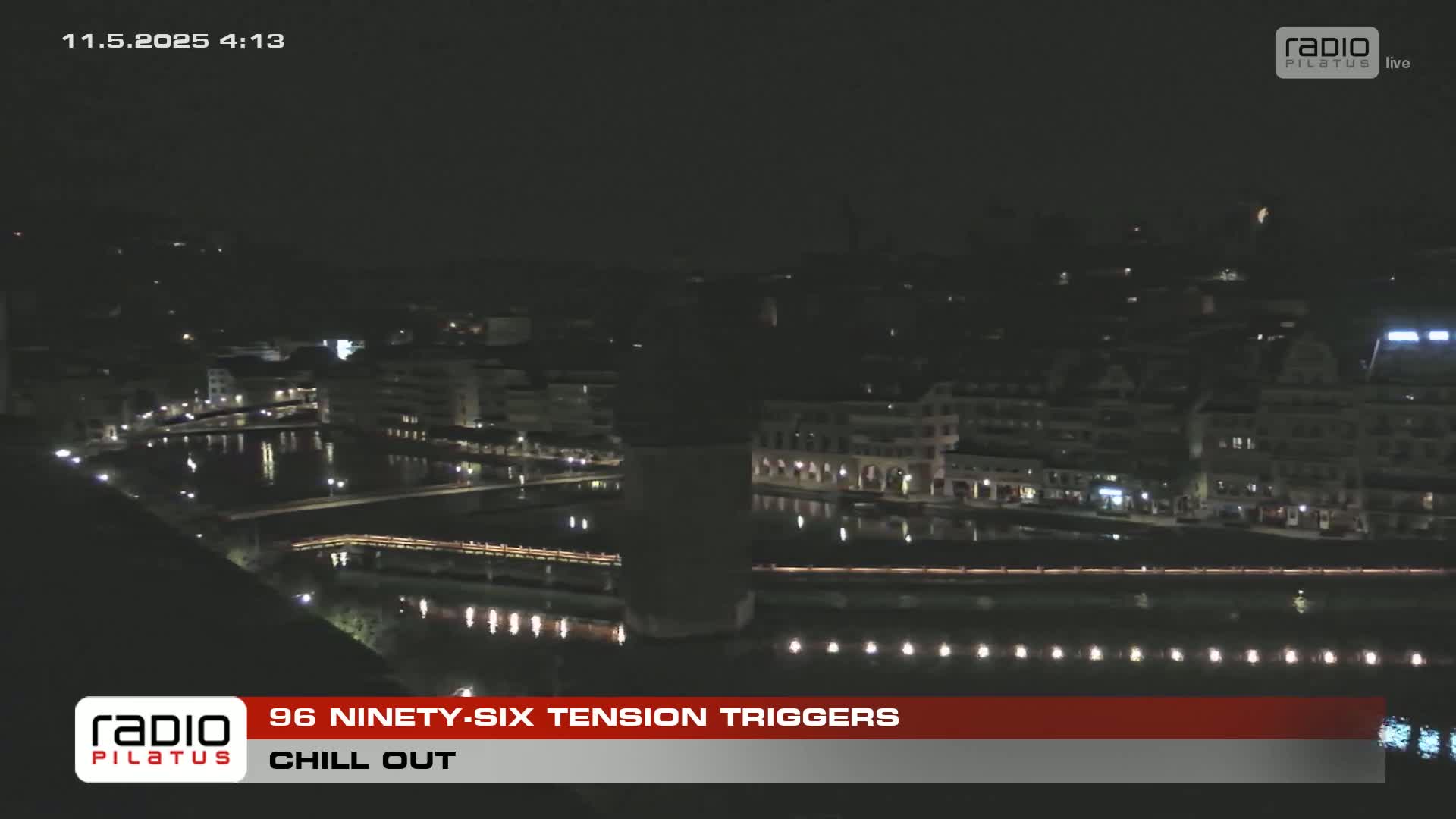 A nighttime aerial view of a city waterfront with calm water reflecting the lights of buildings along the shore.