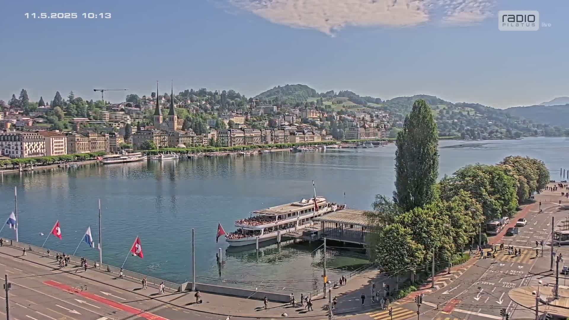 A lakefront city on a sunny day, with a large passenger boat docked near a waterfront road.
