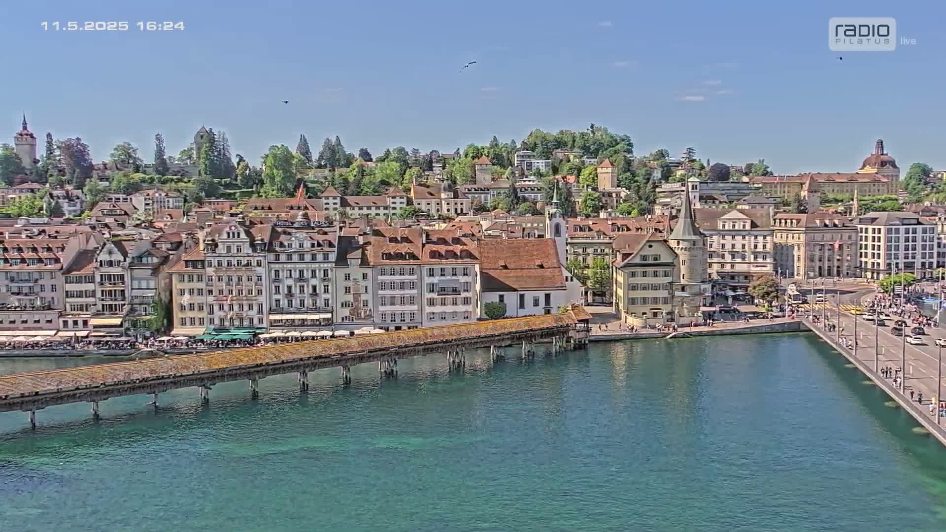 A long wooden bridge spans a teal-colored body of water, connecting to a picturesque town of buildings with red-tiled roofs under a clear, sunny sky.
