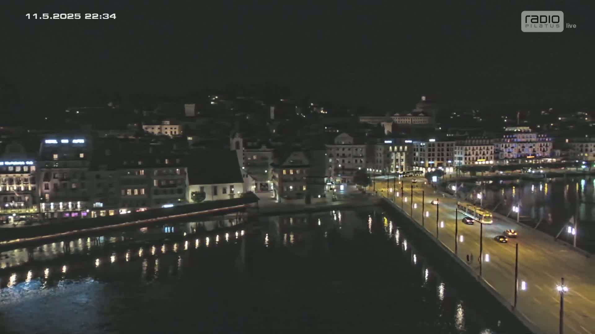 A nighttime aerial view of a city waterfront, showing buildings illuminated along a calm body of water with a bridge and road.