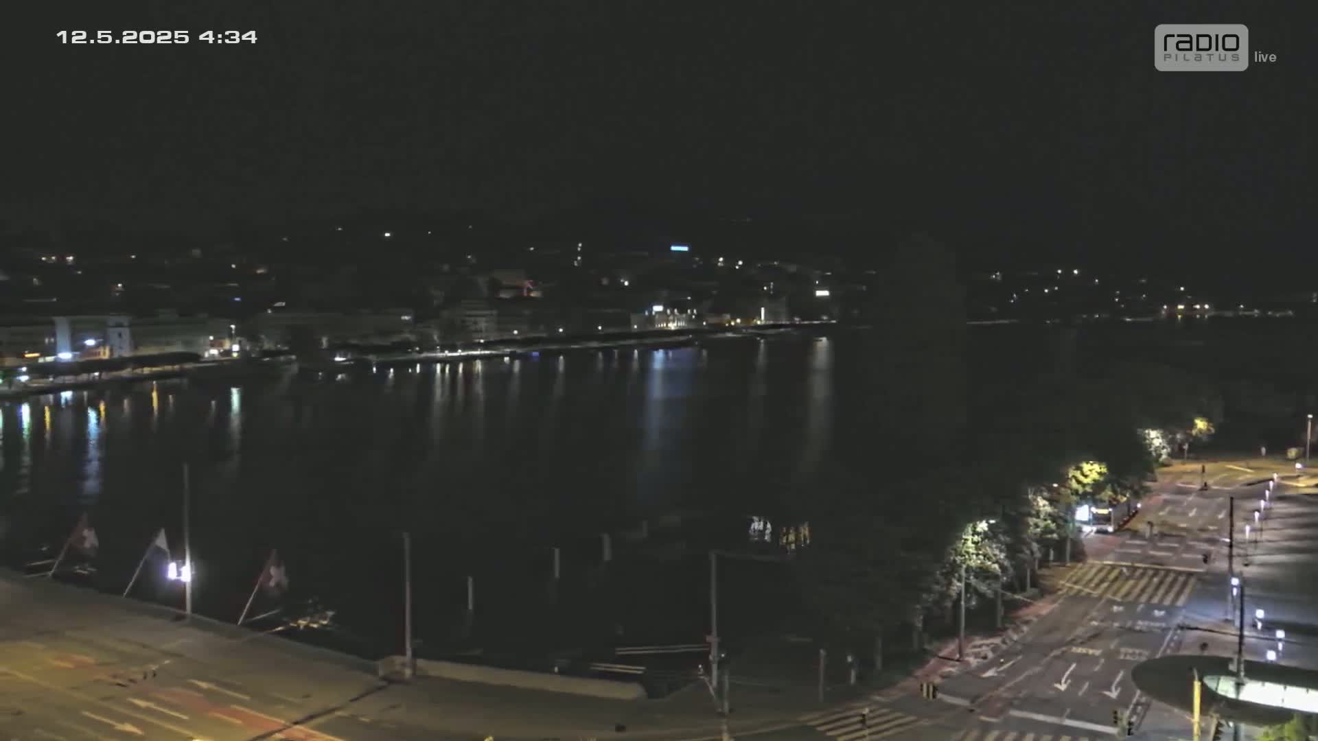 A nighttime view of a calm body of water reflecting city lights, with a road and trees visible in the foreground.