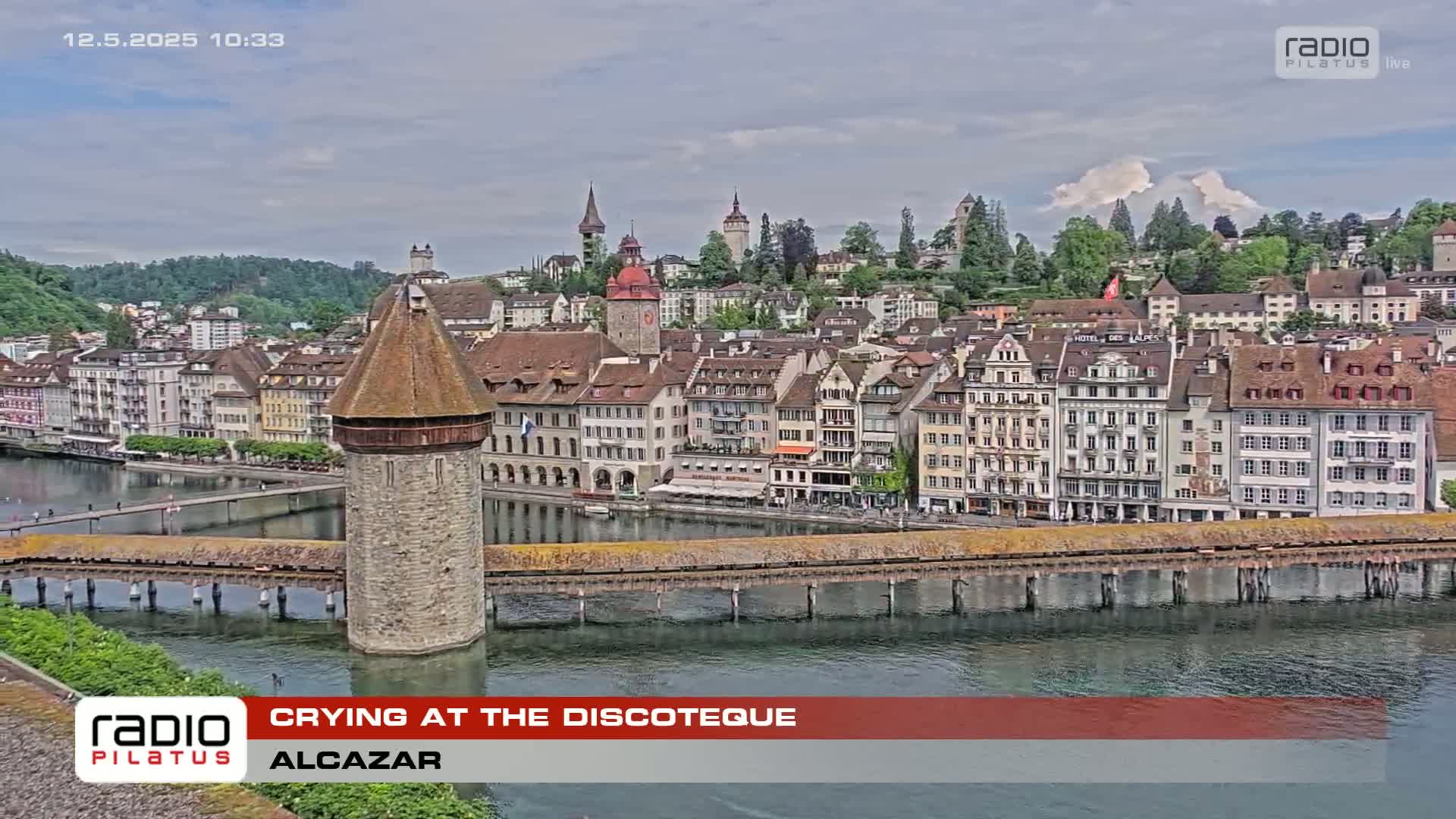 A stone tower sits on a bridge over a calm river, overlooking a picturesque waterfront town under a partly cloudy sky.