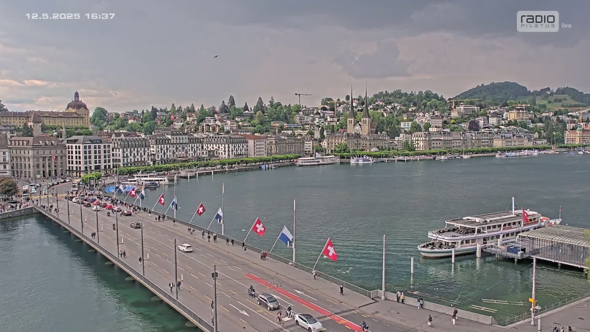 A long bridge with pedestrians and cars spans a body of water, with a city featuring numerous buildings and a docked passenger ship visible in the background under an overcast sky.