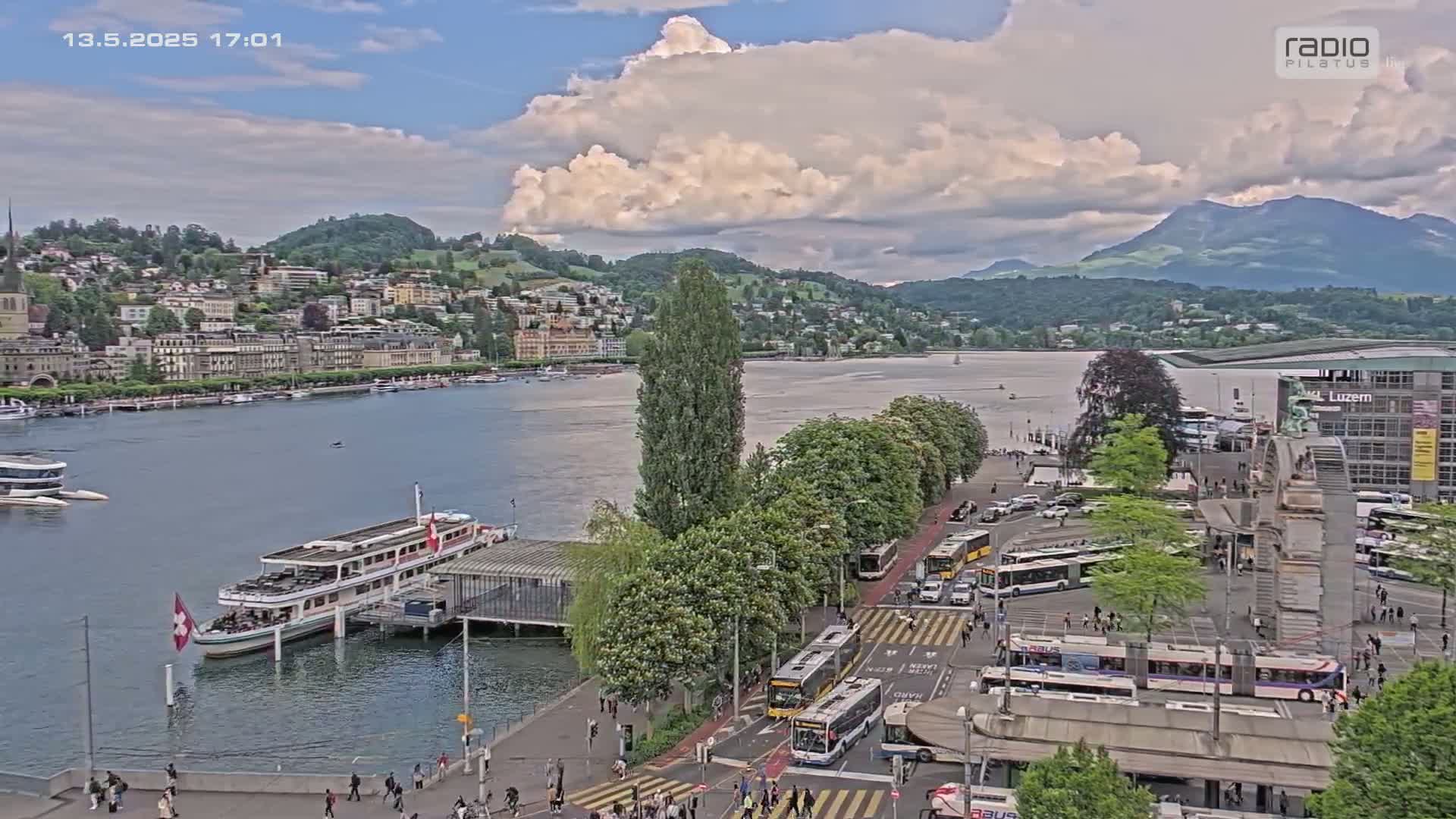 A partly cloudy day overlooking a lakefront city with boats docked, buses at a transit stop, and a mountain range in the background.