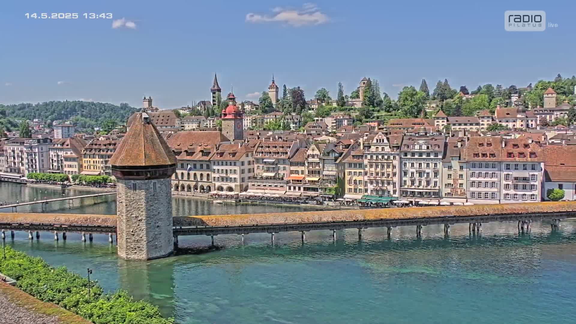 A stone tower on a covered wooden bridge spans a teal river, overlooking a town of multi-storied buildings under a clear blue sky.