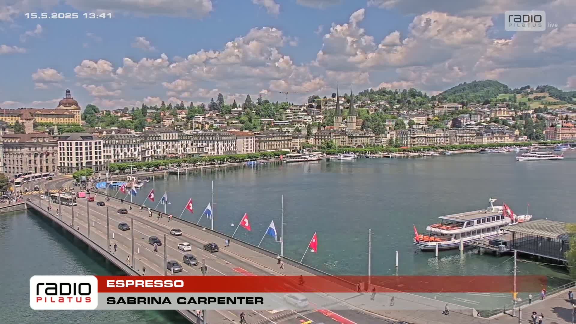 A city waterfront on a partly cloudy day shows a bridge with cars driving across it, boats docked on the water, and buildings lining the shore.