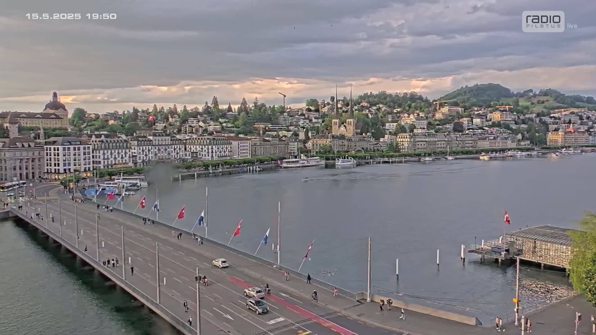 A long bridge with pedestrians and cars crosses a body of water, with a city skyline and overcast skies visible in the background.
