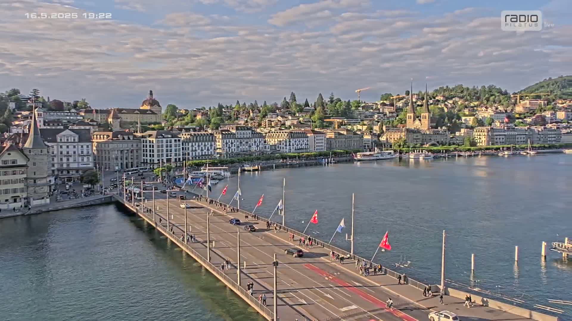 A long bridge spans a body of water, with a city of buildings and a hill in the background under a partly cloudy sky.