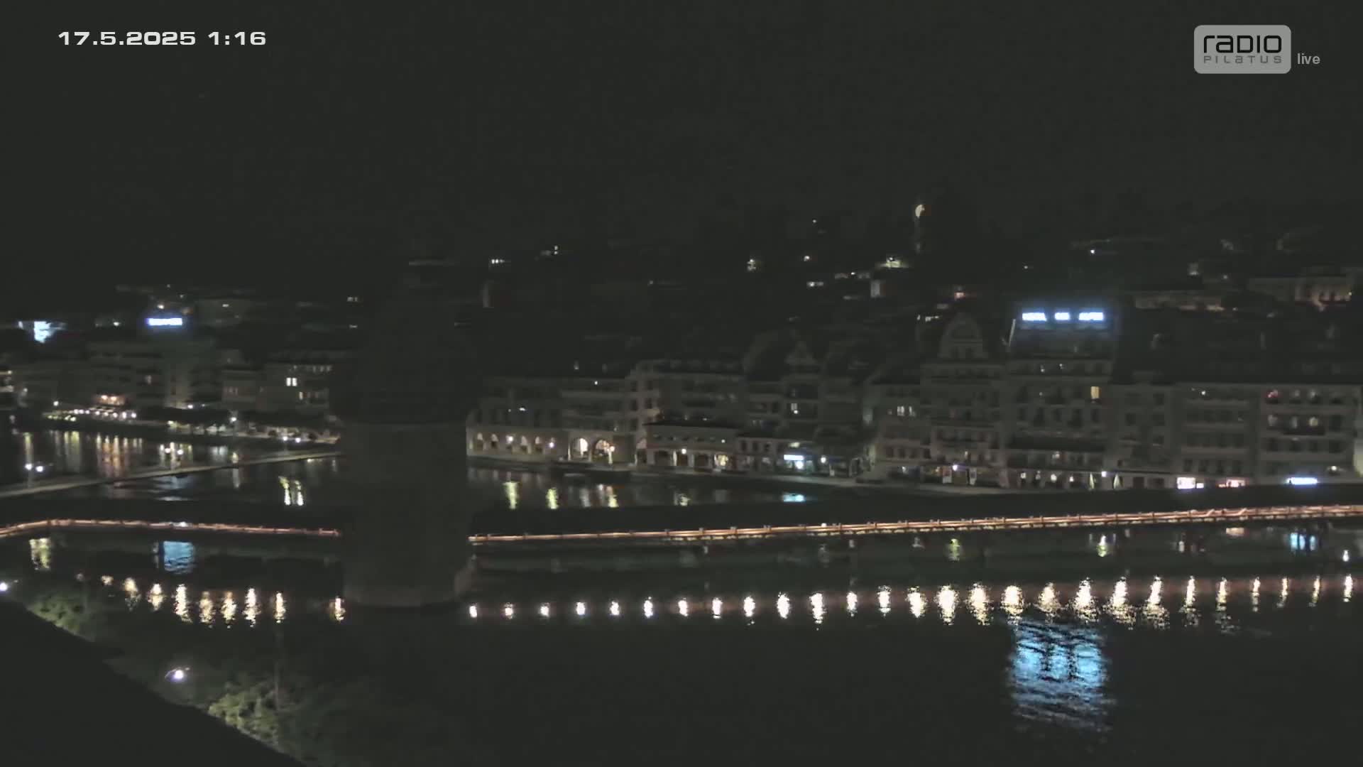 A nighttime view of a city waterfront with buildings and a bridge lit up, reflecting in dark water under a clear sky.