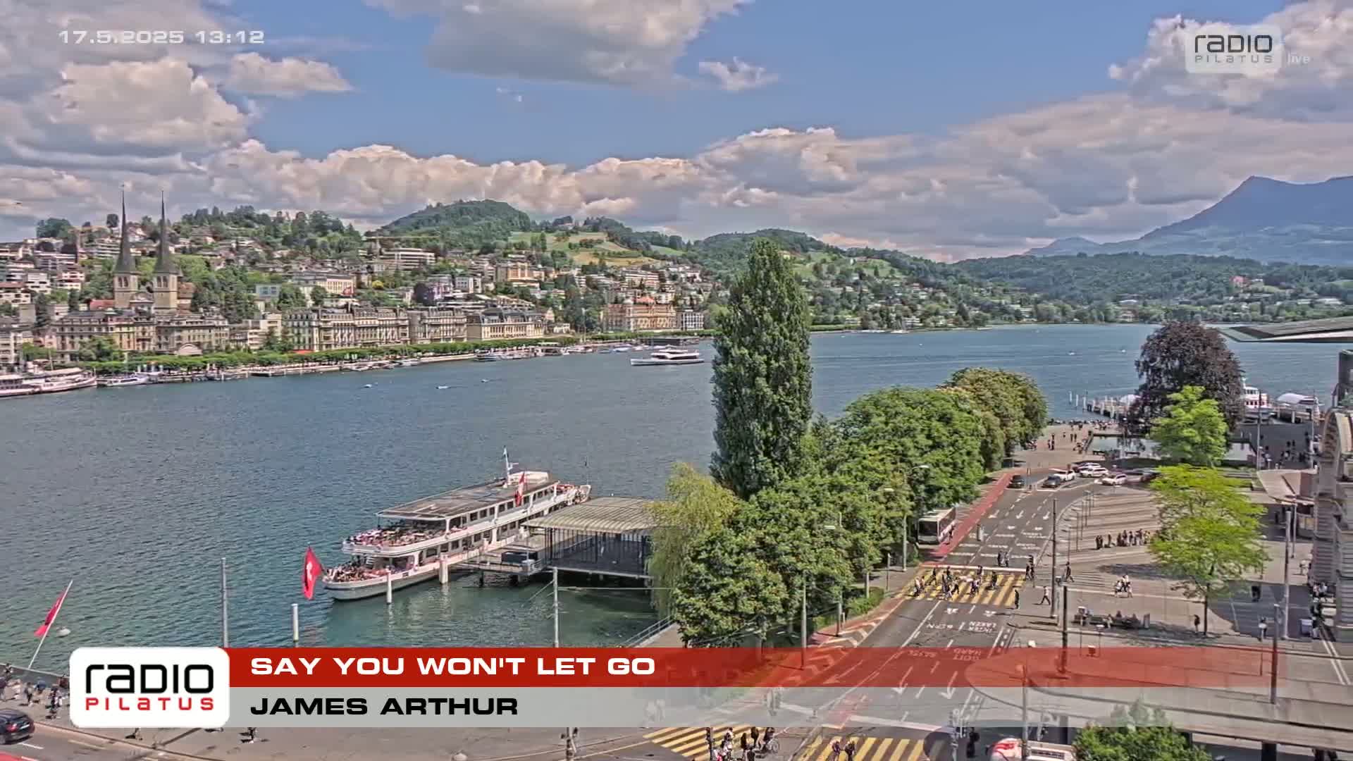 A partly cloudy day shows a lakeside town with a large passenger boat docked near a paved area with pedestrians and vehicles.