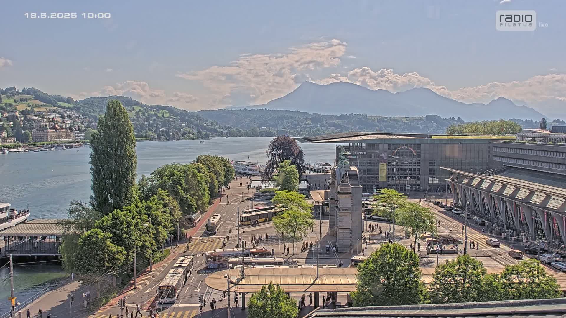 A sunny day reveals a lakeside city scene with a large building, numerous buses and pedestrians, a body of water with boats, and mountains in the distance.