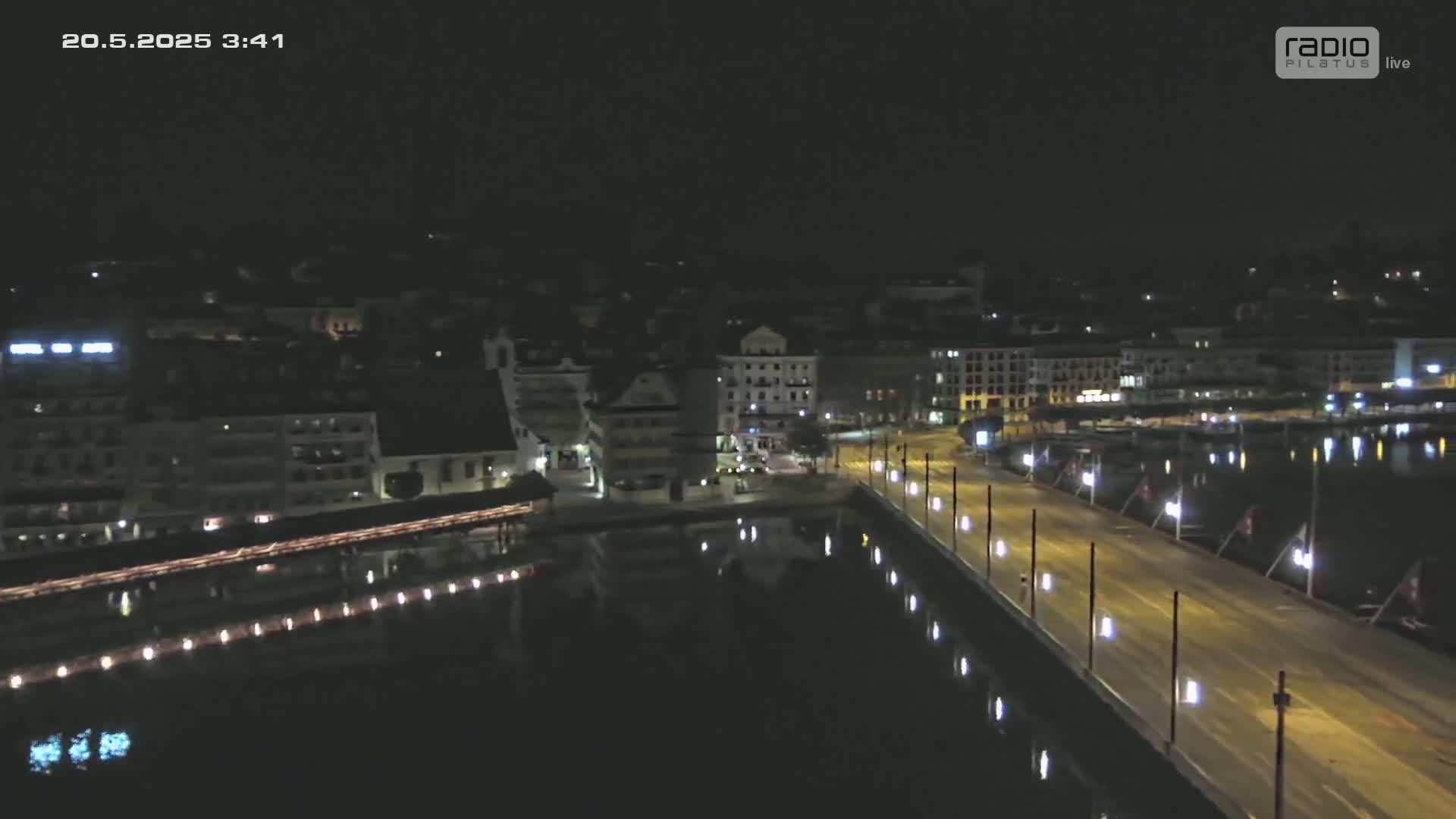 A nighttime aerial view of a city waterfront, showing a bridge and buildings illuminated along a dark body of water under a clear sky.