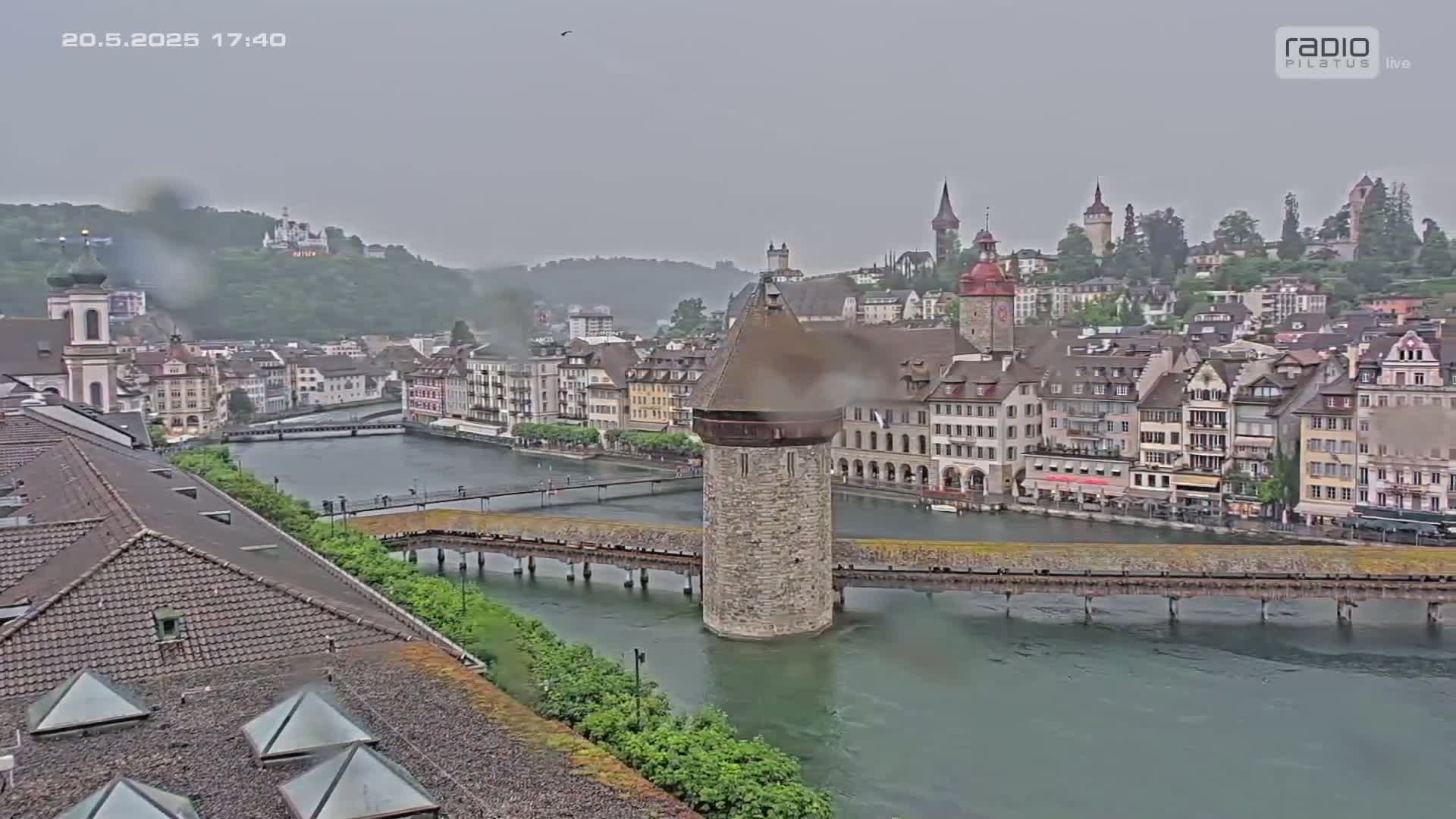 A view of a stone water tower on a bridge spanning a river, with a rain-drenched town and hills in the background under an overcast sky.