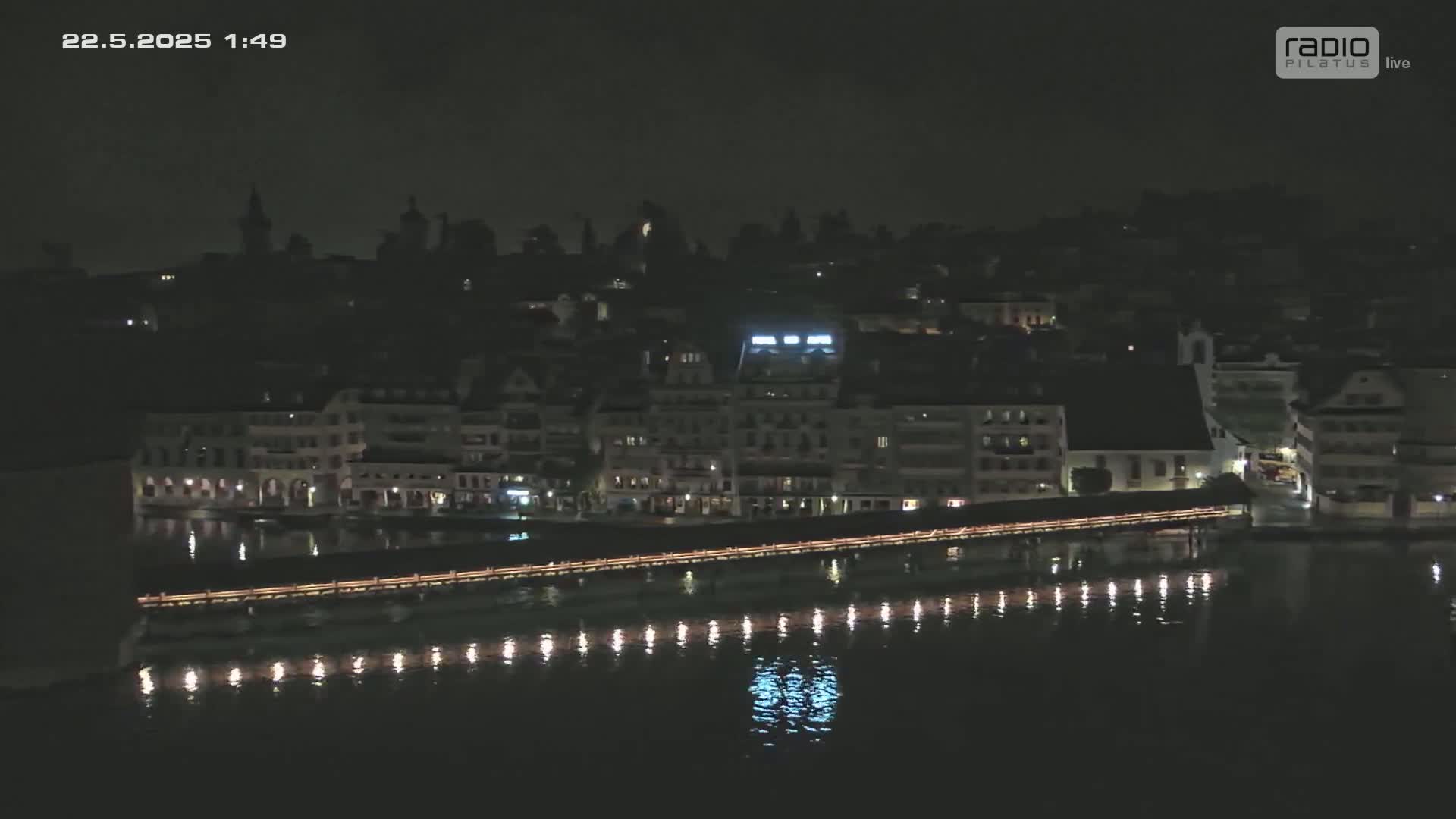 A nighttime aerial view shows a bridge lit with lights spanning a dark body of water, in front of a dark cityscape.