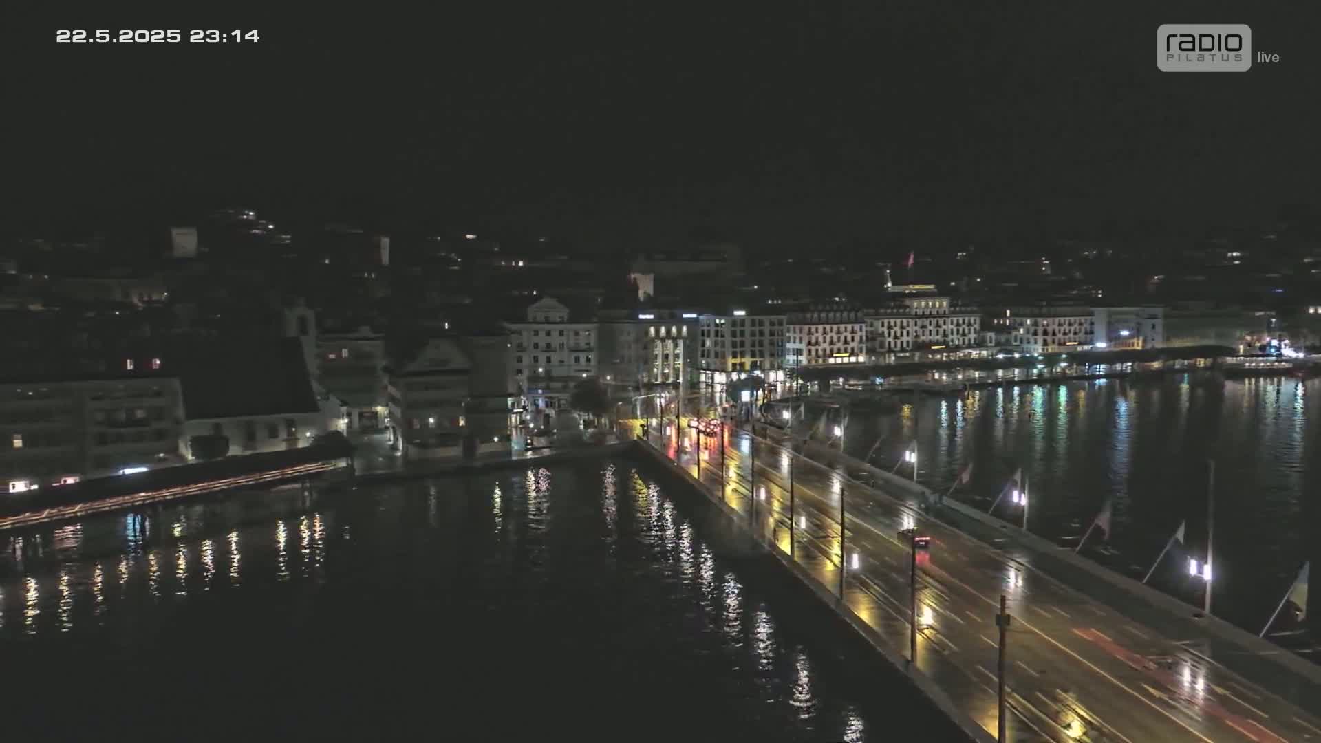A rain-slicked bridge at night, illuminated by streetlights, stretches across a dark body of water reflecting city lights from buildings along the shore.