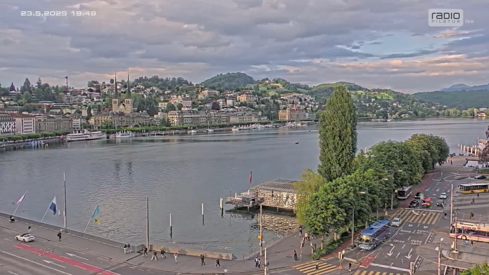 A lakeside city with buildings on a hill, a calm lake with boats, and a roadway with vehicles under a partly cloudy sky.