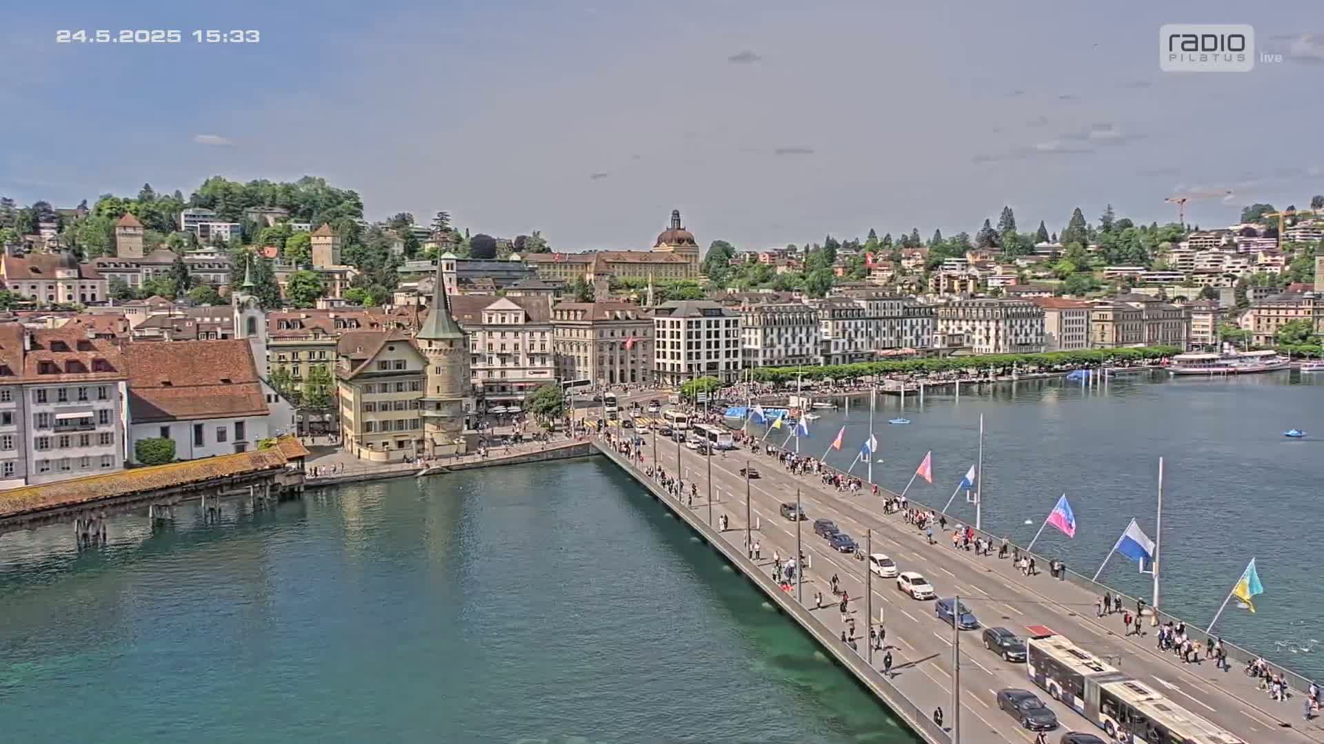 A bridge lined with people stretches across a calm, teal-colored body of water toward a city of European-style buildings on a sunny day.