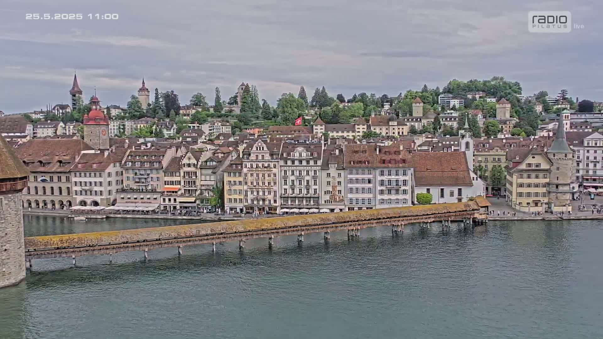 A waterfront European town with many buildings and a covered bridge stretches across a calm river under an overcast sky.