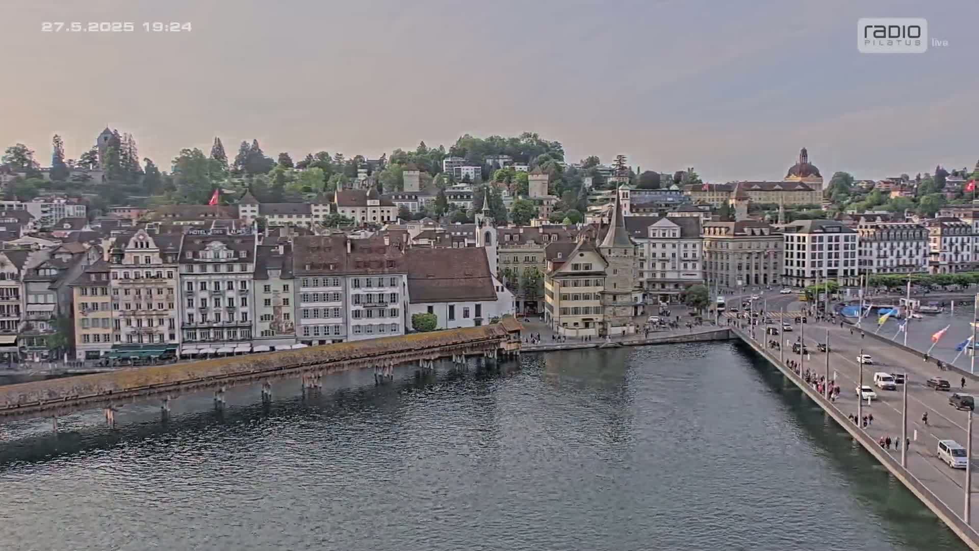A city with many buildings sits on a hill overlooking a river and bridge, under a partly cloudy sky.