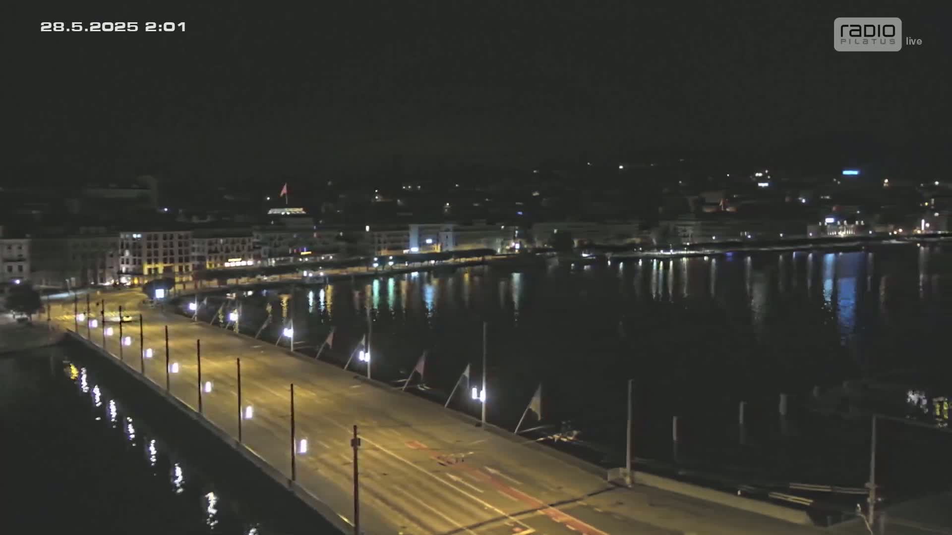 A nighttime aerial view shows a city's waterfront with a bridge extending across calm water, lined with lights.