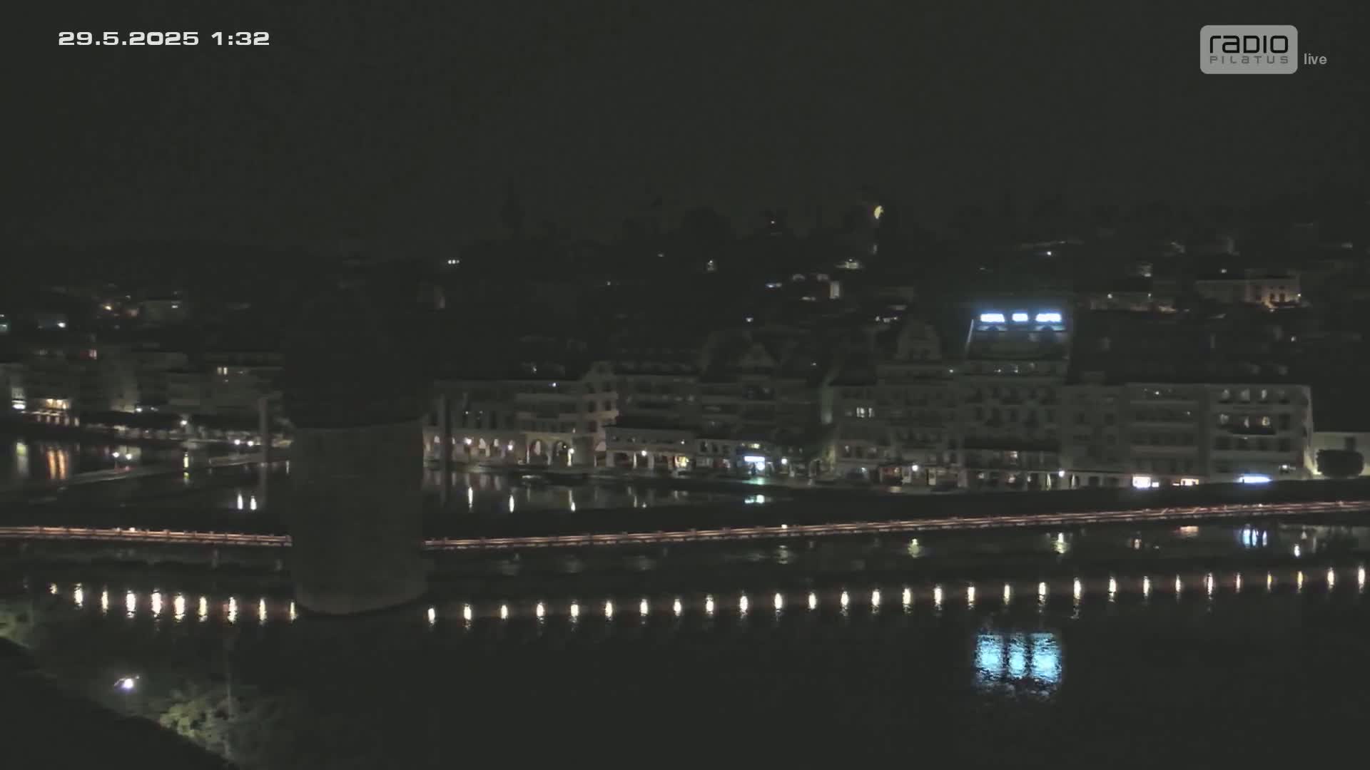 A nighttime view of a city waterfront with buildings illuminated along a calm body of water, under a dark sky.