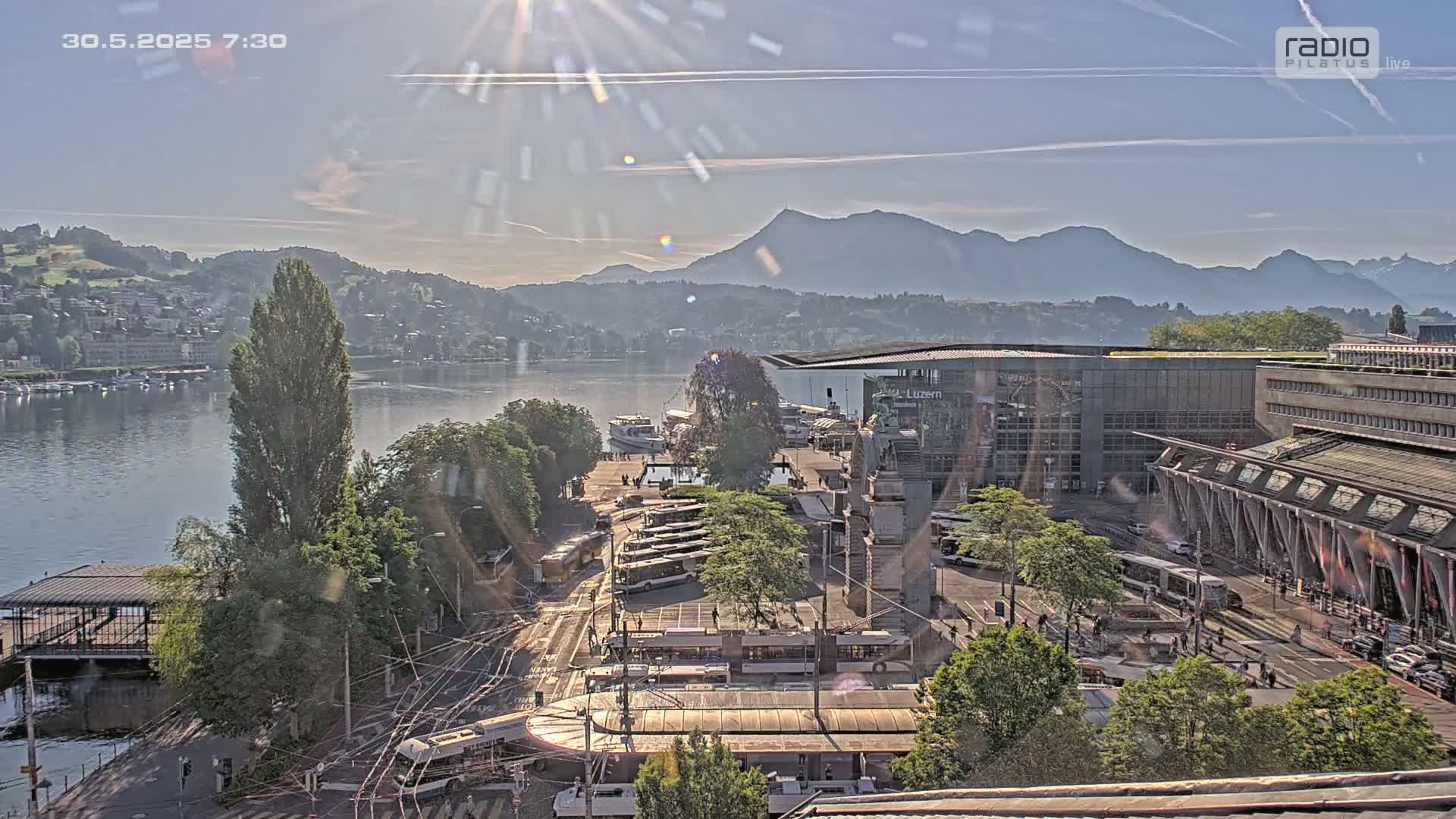 A sunny day overlooking a lake with boats, a city, mountains in the background, and a plaza with several buses and trams.