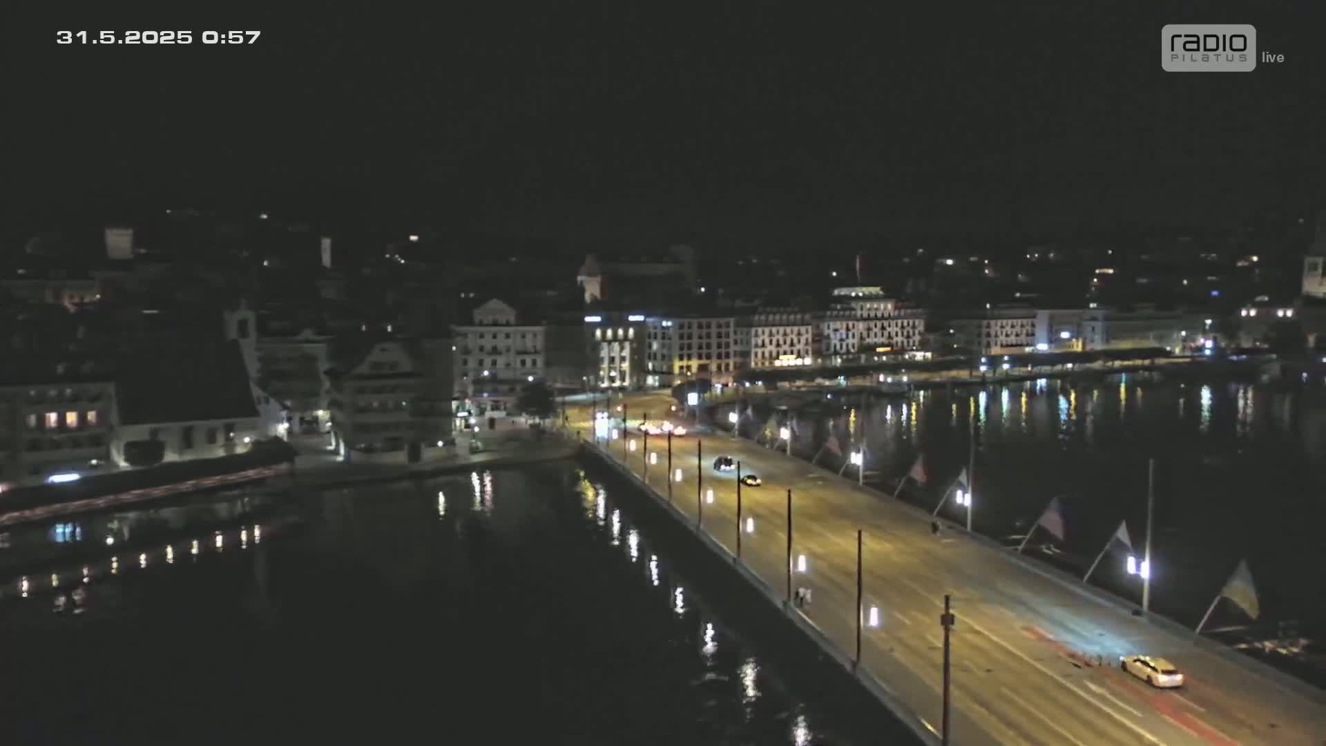 A nighttime, long shot of a bridge over water with several cars driving on it, and buildings lining the waterfront.