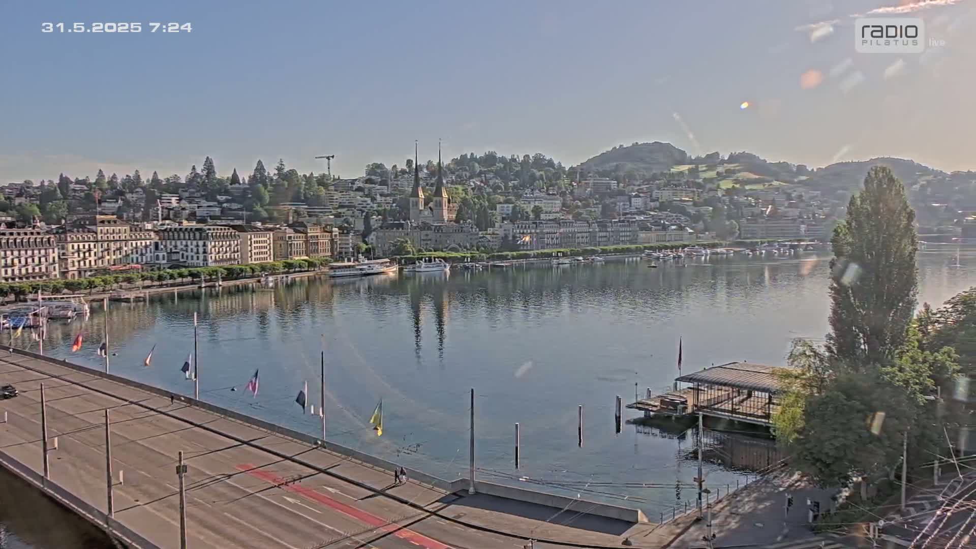 A calm lake reflects a city skyline on a sunny morning, with a bridge and roadway visible in the foreground.