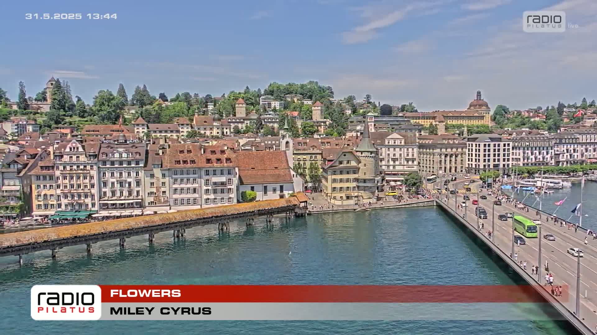A sunny day overlooking a European city with a bridge crossing a body of water, showing numerous buildings, pedestrians, and vehicles.