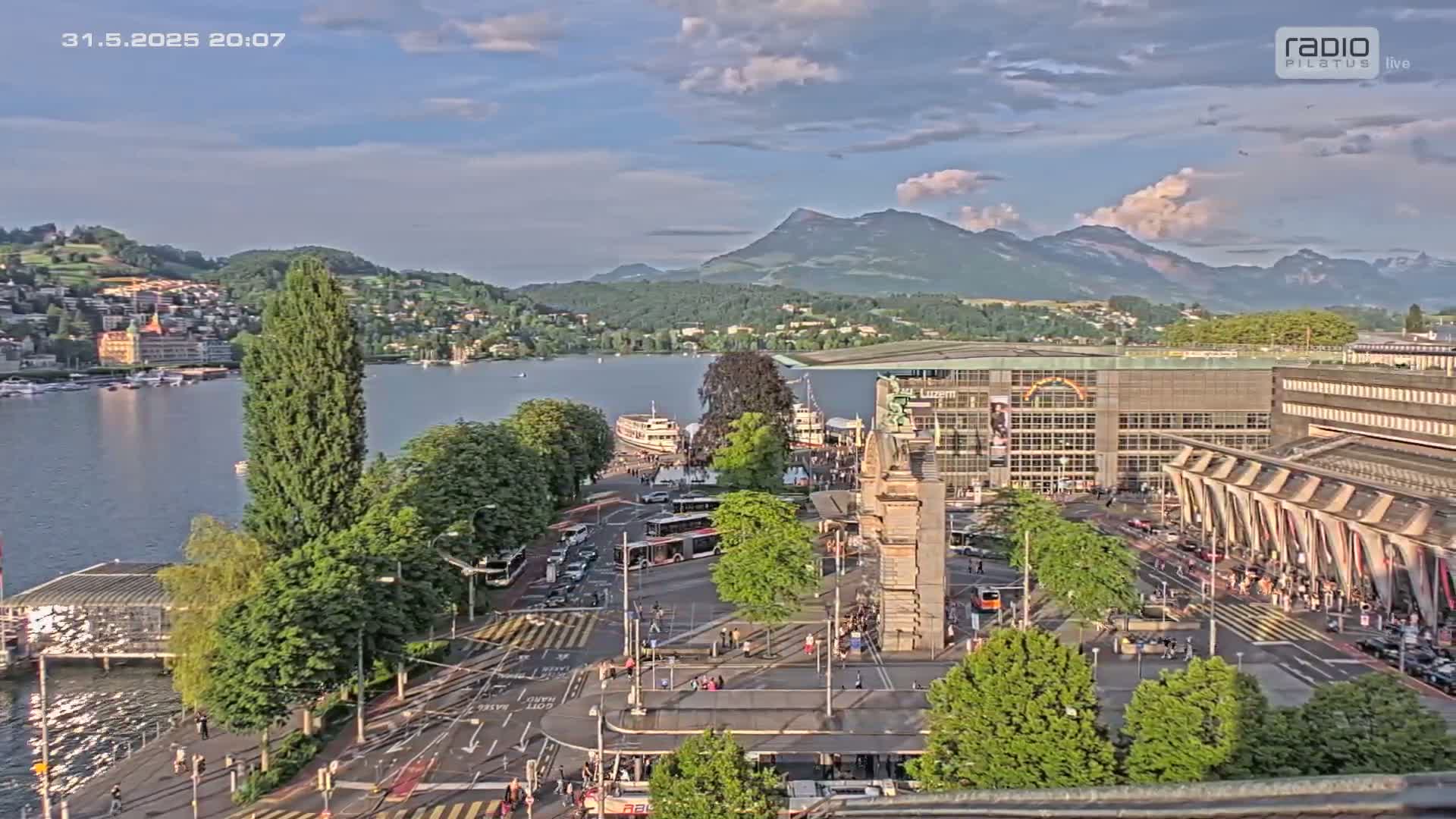 An elevated view shows a city square near a lake, with boats on the water, mountains in the background, and partly cloudy skies.