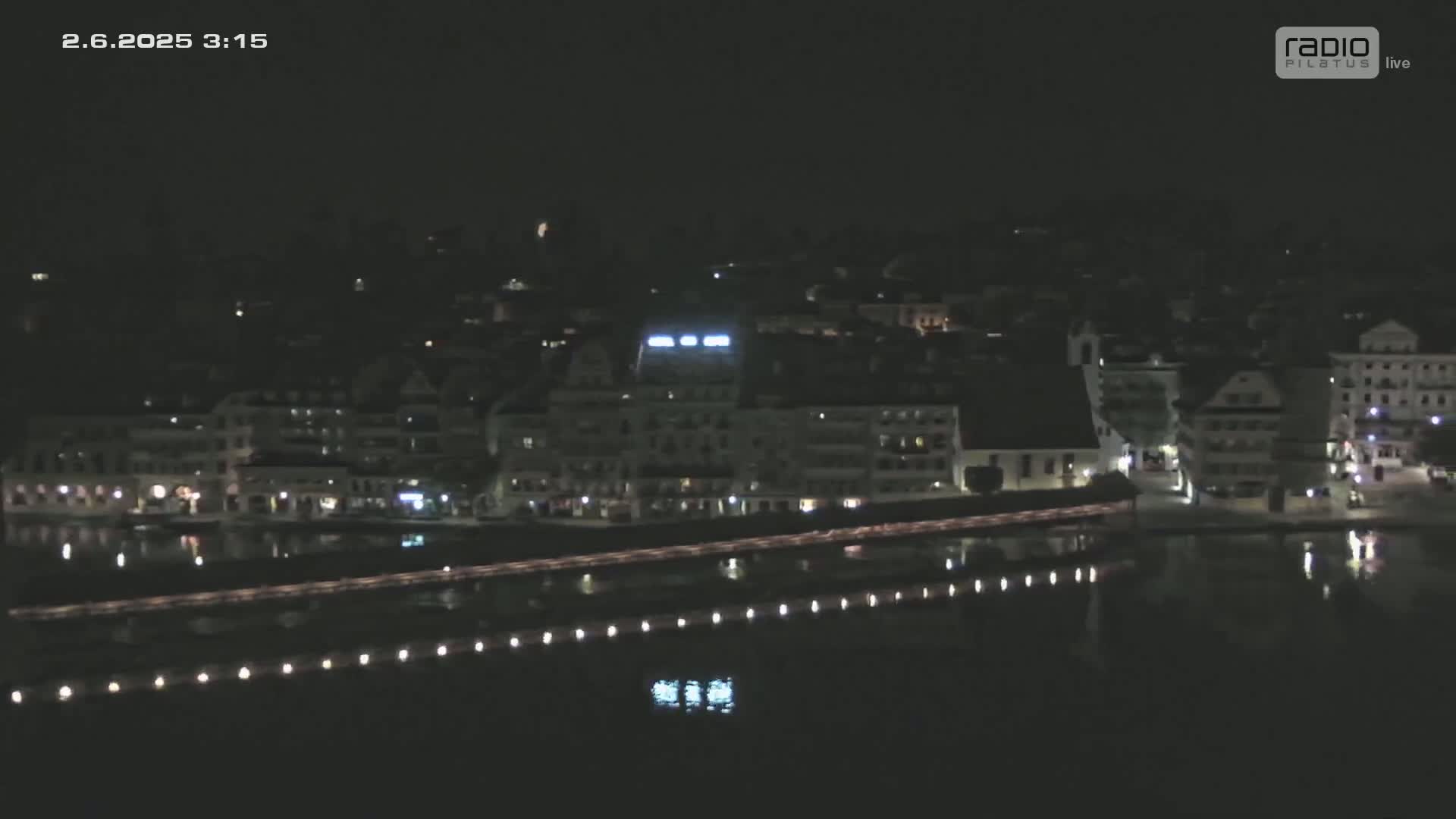 A nighttime, high-angle view shows a waterfront town with buildings and lights reflecting in dark water, under a clear, dark sky.