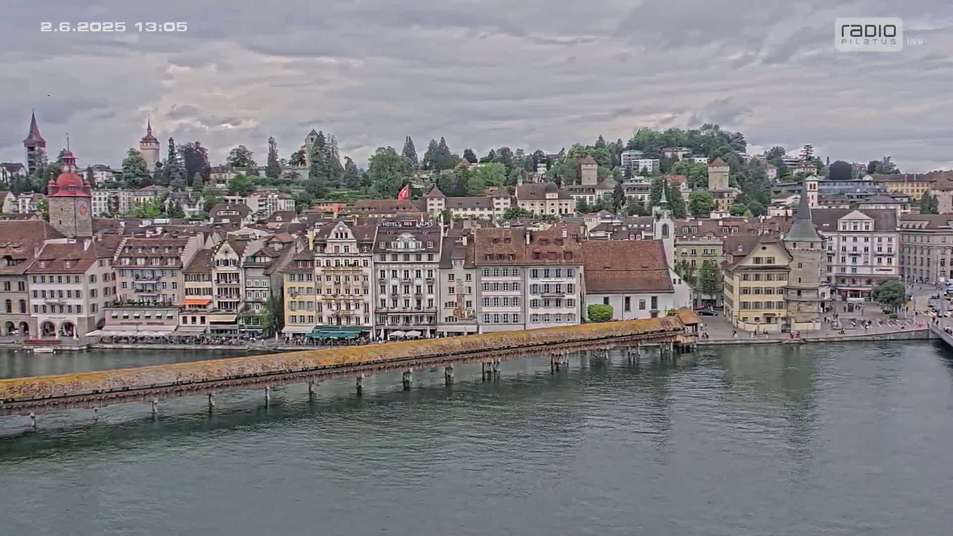 A long wooden bridge extends over calm water towards a picturesque European city under a cloudy sky.