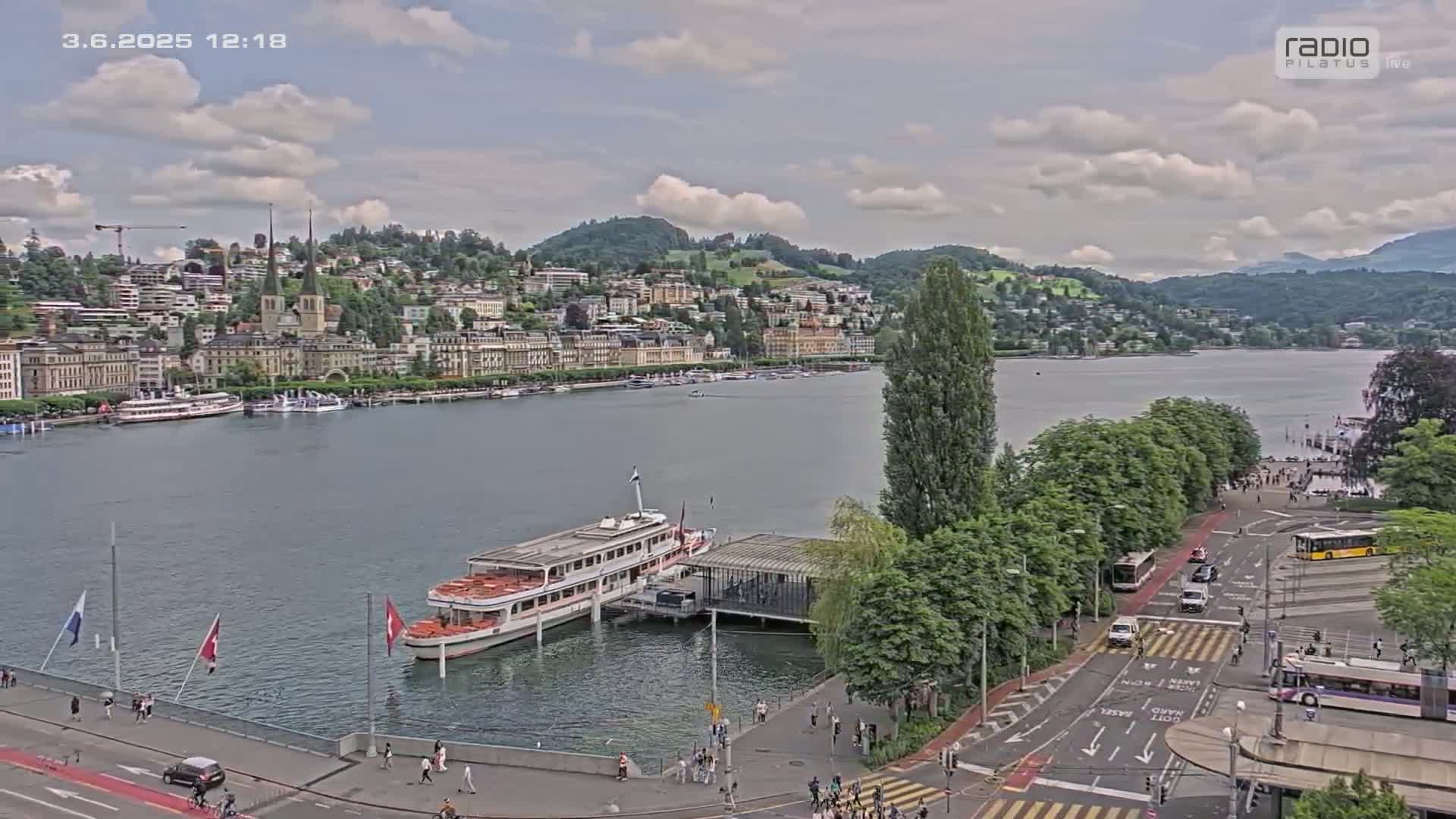 A partly cloudy day reveals a lakeside town with a docked passenger ship,  surrounded by trees and roads with several vehicles and pedestrians.