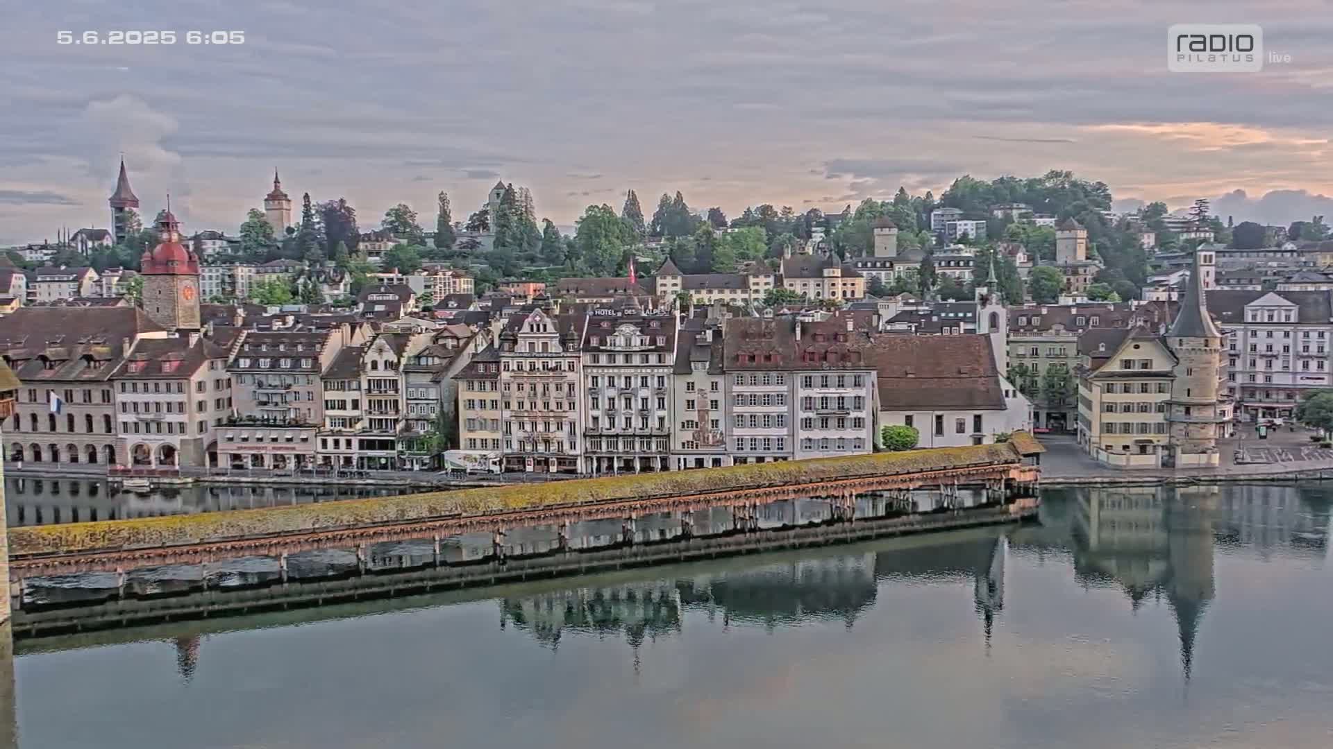 A picturesque European town sits beside a calm river under a cloudy sky, with its buildings and a covered bridge reflecting in the water.