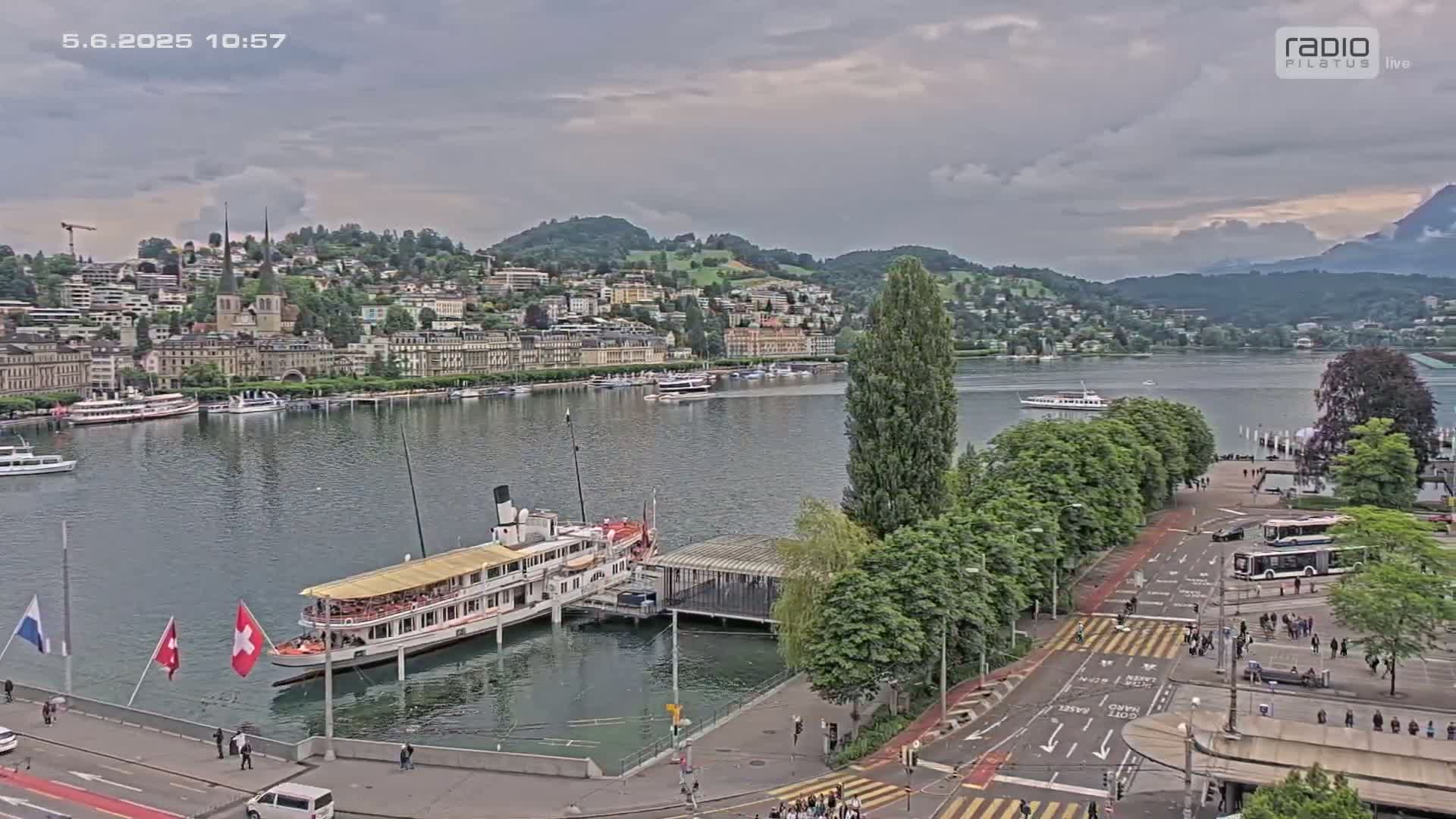 An overcast day reveals a paddle steamer docked near a lakeside town, with calm water, lush greenery, and a pedestrian area.