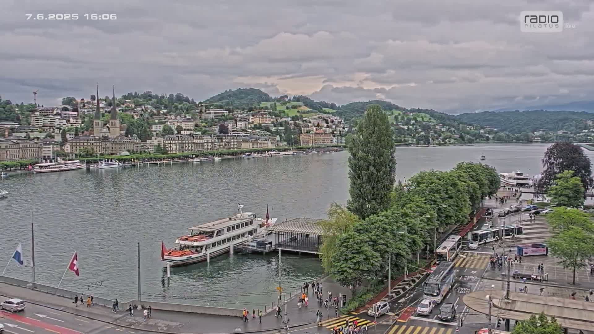 A lakefront city under a cloudy sky, with boats docked near a shoreline featuring buildings, trees, and people walking along a roadway.