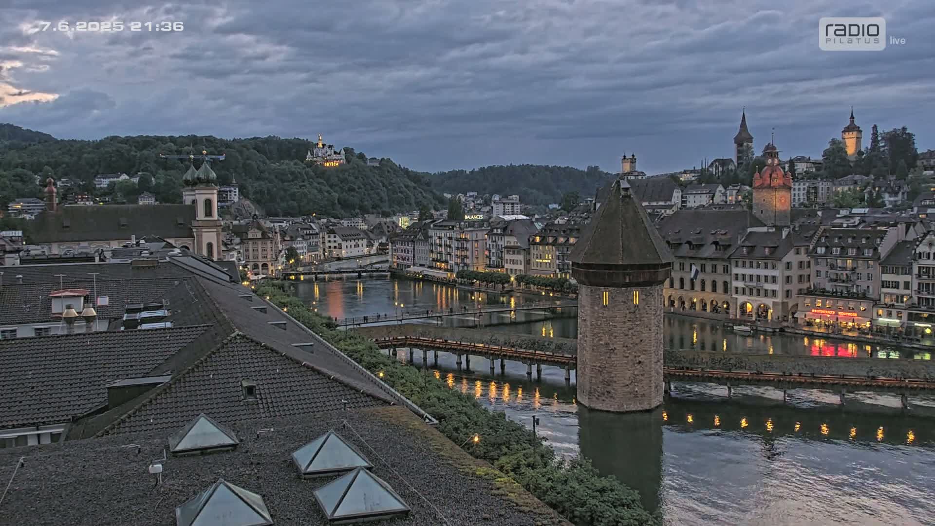 A twilight view of a European city with a river running through it, featuring a stone tower on a bridge, and many buildings with lit windows under a cloudy sky.