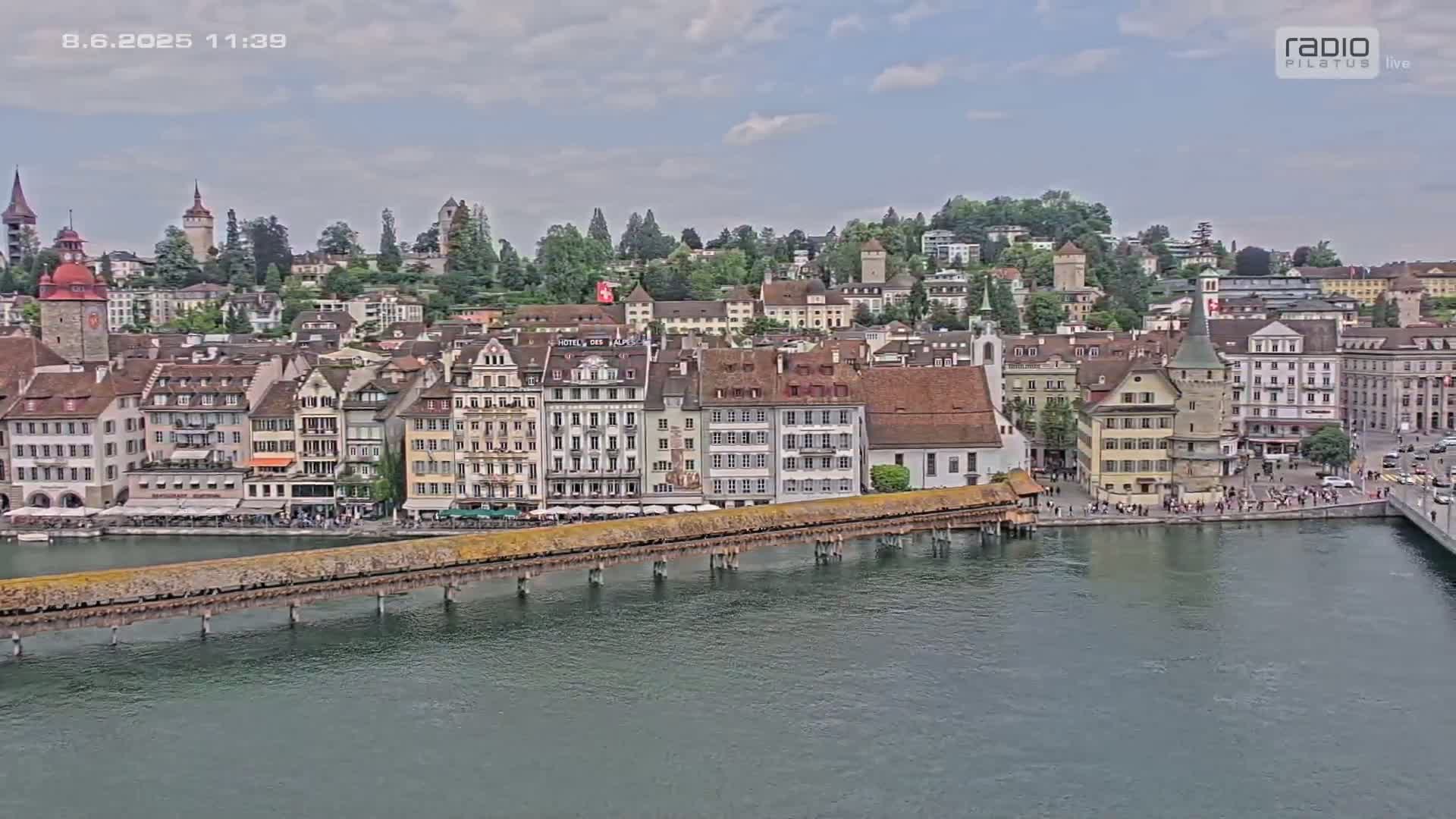 A picturesque waterfront town with numerous buildings, a long covered bridge, and a calm river under a partly cloudy sky.